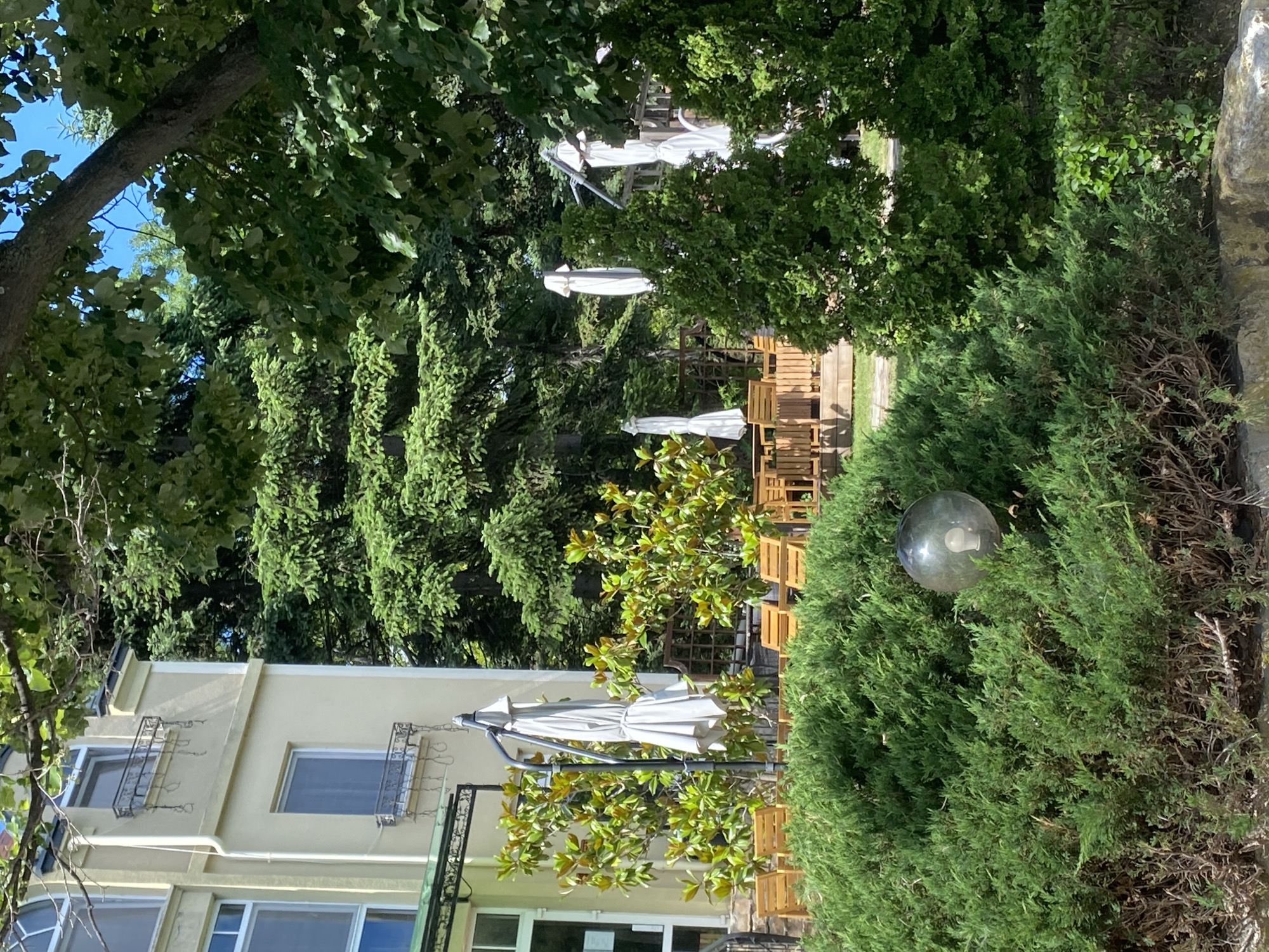 The image depicts an outdoor seating area with lush greenery, suggesting a pleasant day, likely in the morning or early afternoon, given the bright sunlight filtering through the leaves. The scene is set in Varna, Bulgaria.
Dominating the foreground is a dense, green evergreen bush, likely a juniper, with a round, translucent glass orb hanging from it, possibly a decorative garden light or ornament. Behind this bush, a pale yellow building with multiple windows is visible. The building features decorative wrought iron window grates on the upper floor and appears to be a part of a restaurant or hotel.
Further back, a collection of outdoor furniture, including several wooden chairs and tables, are arranged under large, white patio umbrellas. These umbrellas are partially folded, suggesting they might be closed or preparing to be opened. The furniture and umbrellas are interspersed with various trees and shrubs, creating a shaded and inviting atmosphere.
The overall impression is one of a well-maintained, tranquil outdoor space, possibly a courtyard or garden area attached to a building. The bright sunlight and abundant foliage indicate a warm, possibly summer day. No people are visible in the image. There is no visible text in the image.