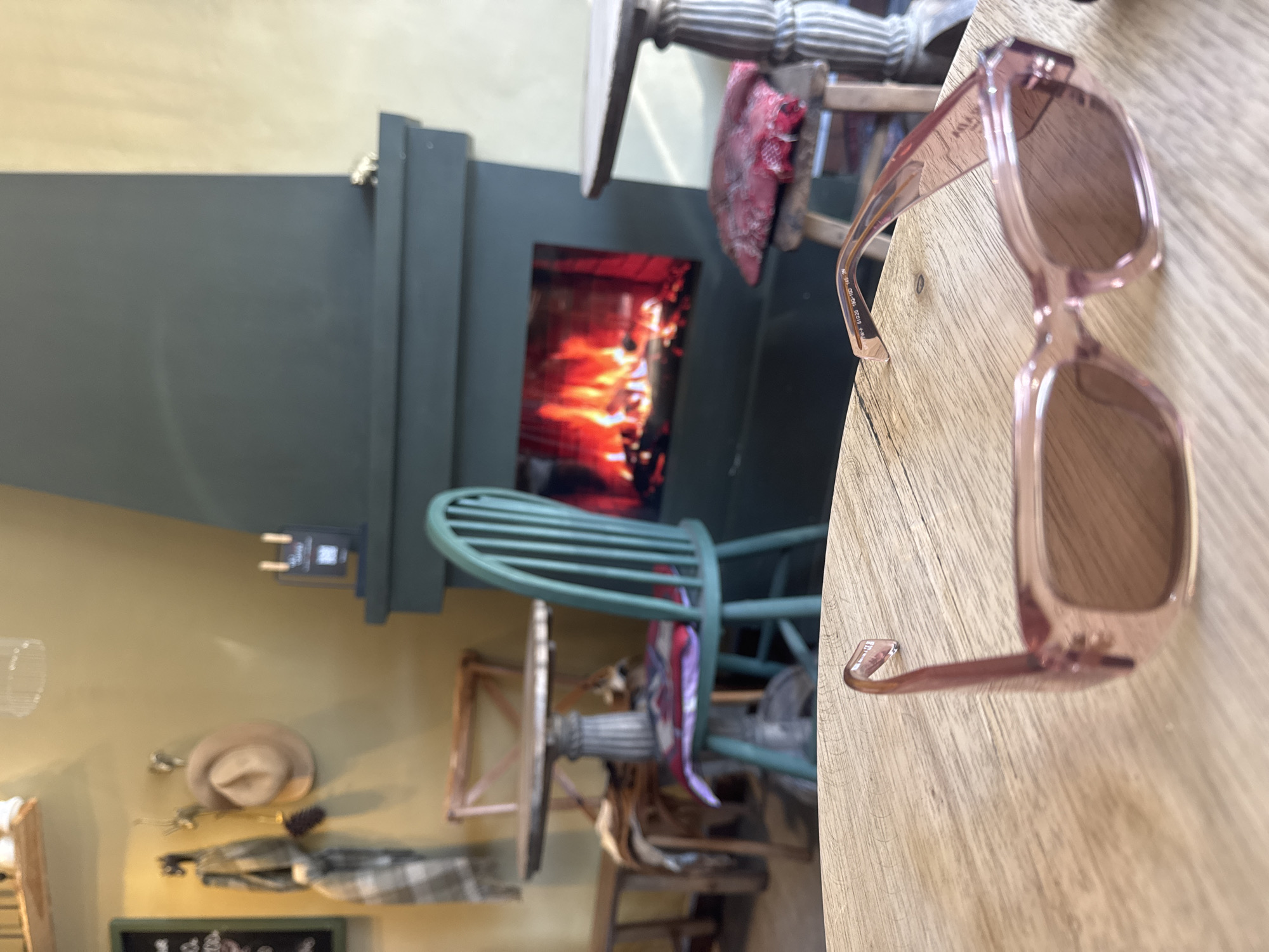 The image captures an indoor scene, likely a restaurant interior, featuring a fireplace with visible flames, a green chair, wooden furniture, and a pair of pink sunglasses placed on a wooden table. A hat and coat are hanging on the wall. The location in the city is unknown.