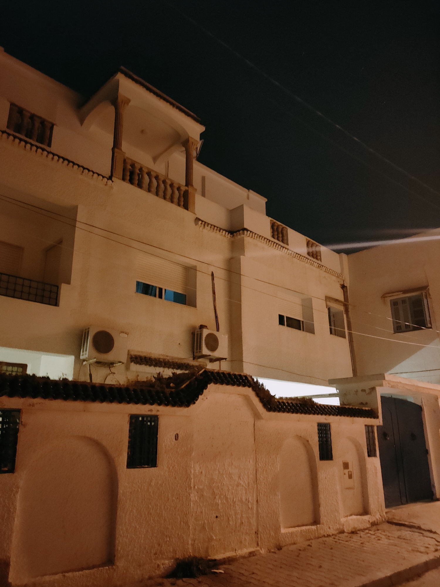 The image shows a facade of a building in Tunis, Tunisia, at night. The building is predominantly white with some terracotta-colored accents, particularly on the roof tiles and decorative elements. The architecture features a balcony with decorative columns and a balustrade. Several windows are visible, some with closed shutters and others with blinds. Two air conditioning units are mounted on the exterior wall.
In the foreground, there is a low, textured white wall with arched niches and small, barred windows. The wall is topped with dark, tiled coping. A dark, large double door is visible to the right of the frame, suggesting an entrance or garage. The ground in front of the wall is paved with cobblestones. The sky above is dark, indicating nighttime, with a few faint specks of light that could be stars or distant lights. There are also some power lines visible against the night sky. The overall impression is of a residential building in a somewhat aged but well-maintained state.