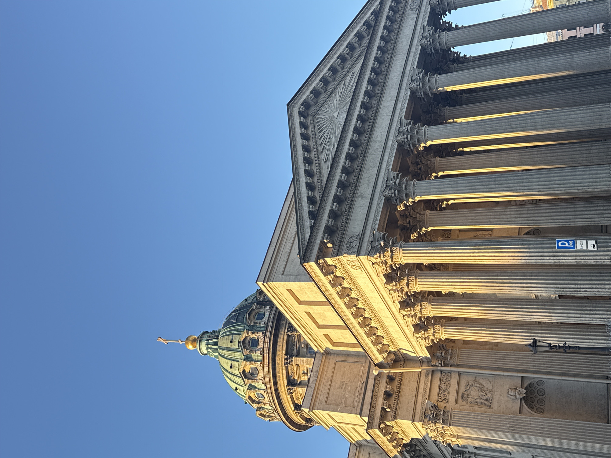 The image shows a low-angle view of the Kazan Cathedral in Saint Petersburg, Russia, against a clear blue sky. The architectural details of the cathedral are prominent, including the dome, colonnade, and decorative elements. The columns are illuminated by sunlight. A parking sign is visible on the side of the building.