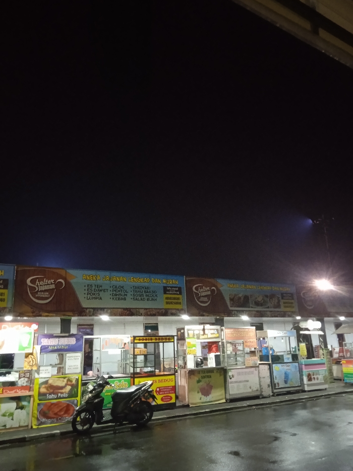 This nighttime image depicts a row of illuminated food stalls, likely a street food market or food court, in Kecamatan Kartasura, Indonesia. The sky above is dark, indicating late evening or night, and the wet, reflective ground suggests recent rain.
The scene is dominated by several vendor kiosks arranged under a long, brightly lit overhead banner. The banner prominently features a "Shelter Jajanan" logo and advertises "ANEKA JAJANAN LENGKAP DAN MURAH" (Variety of Complete and Cheap Snacks). The listed items include "ES TEH," "CILOK," "TAKOYAKI," "ES DAWET," "PENTOL," "TAHU BAKSO," "POKIS," "DIMSUM," "SOSIS BEDUG," "LUMPIA," "KEBAB," and "SALAD BUAH."
Individual stalls below the main banner showcase their offerings. On the left, one stall advertises "TAHU PETIS" (Tofu with shrimp paste sauce) from "Mak Ni," and "Pisang Crispy" (Crispy Banana). A black motorcycle is parked in front of these stalls. Further to the right, other kiosks are visible with signs such as "ES BEDUG" and "MY DAY." Another partially visible sign likely says "KEBAB." The stalls vary in design, some featuring glass display cases and others with colorful graphics.
No people are clearly visible in the image, contributing to a quiet atmosphere despite the vibrant artificial lighting. The bright lights from the stalls and a distant streetlamp on the far right cut through the darkness, illuminating the food vendor area, which appears ready for business.