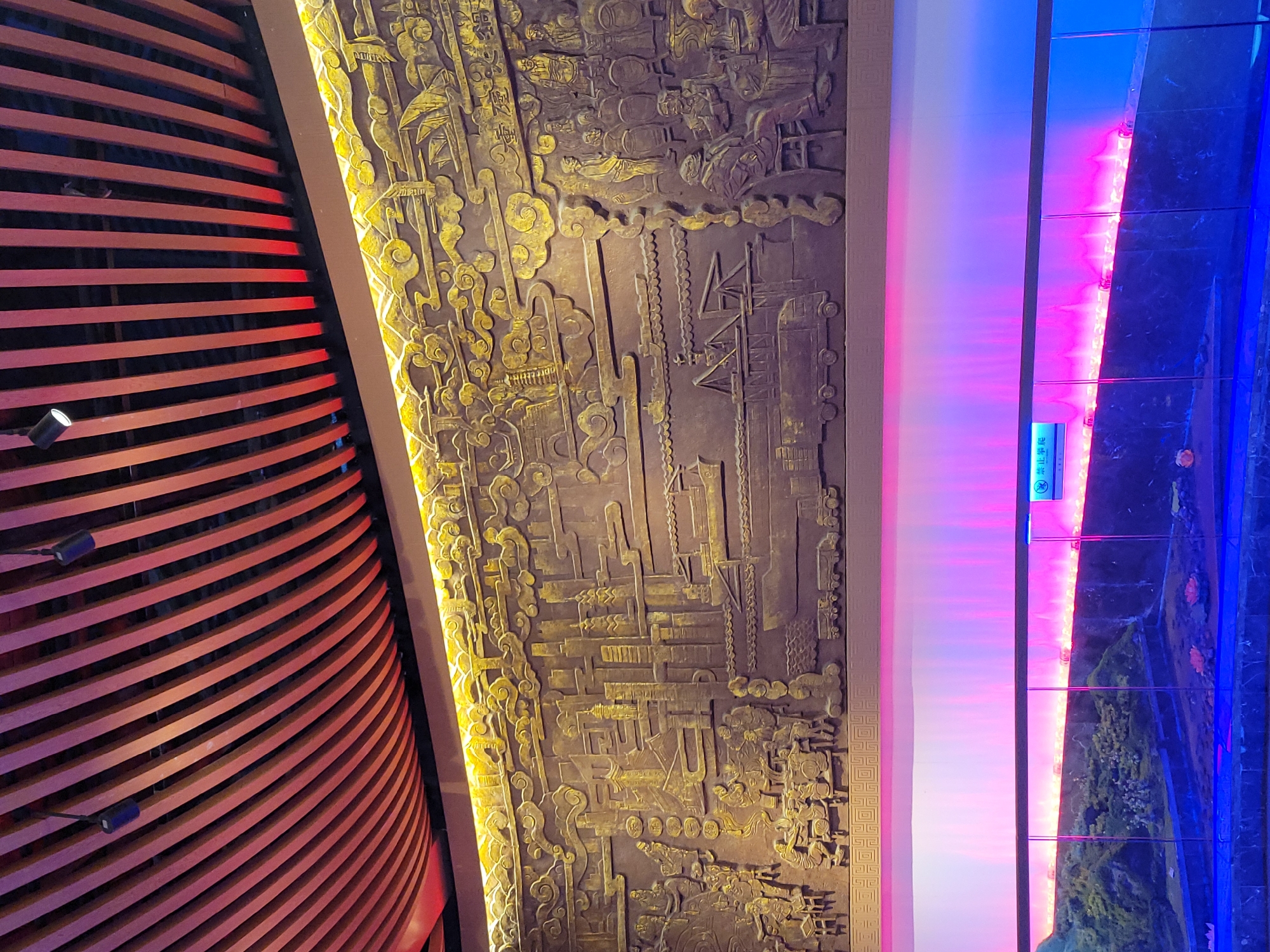 The image captures an interior scene in Hechuan, China. A curved wall of horizontal red-lit wooden slats dominates the left side of the frame. Adjacent to this is a wall with a bas-relief depicting an ancient Chinese scene, featuring figures, landscapes, and architecture in a bronze color. To the right of the bas-relief wall, a vertical section displays a spectrum of colors, transitioning from white to pink and then to blue. This section suggests illuminated glass panels or a similar decorative element. A sign with Chinese characters is visible on the panel. The image exhibits a blend of traditional artistic elements with modern lighting and design.