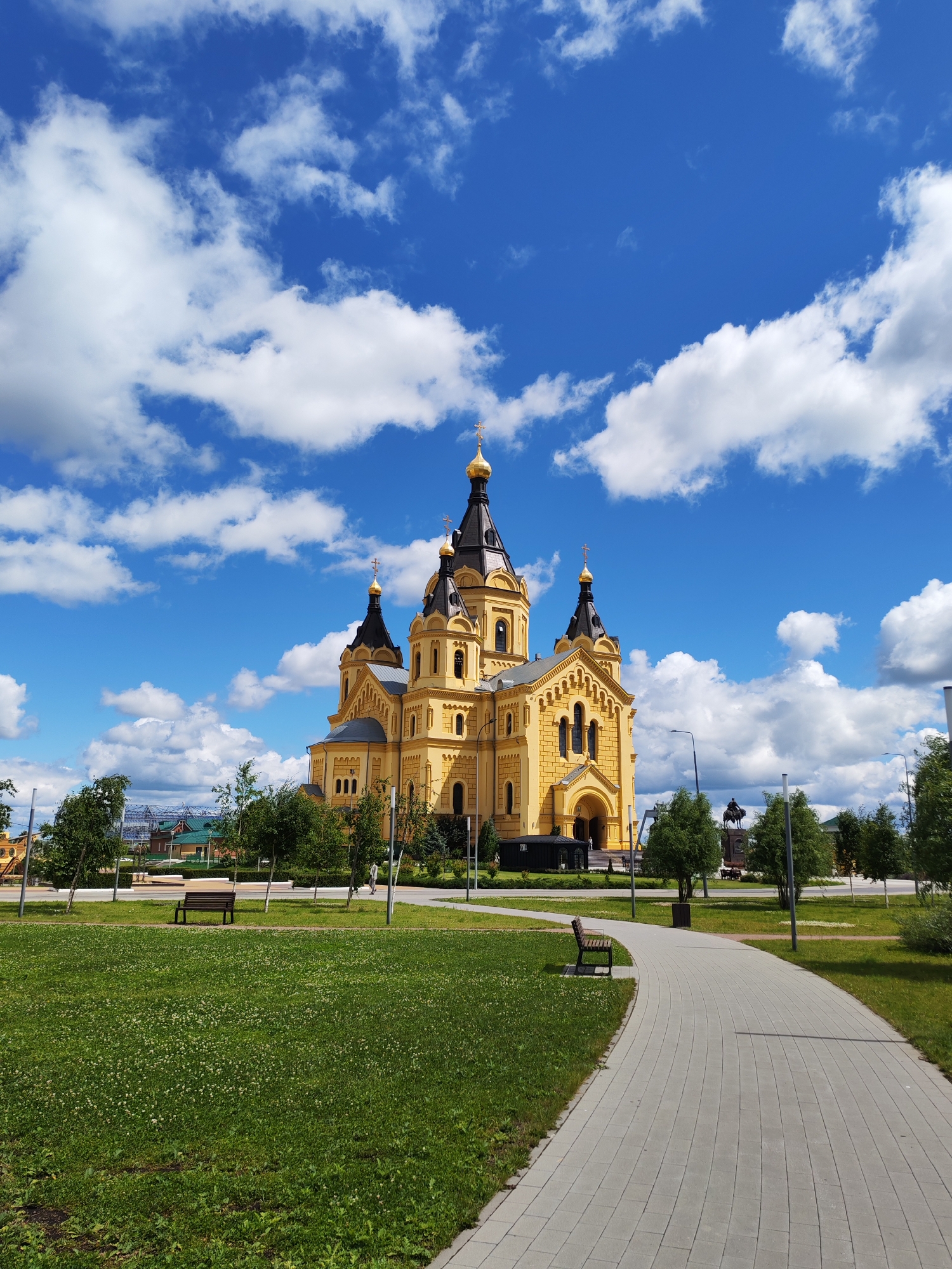 This media file captures a bright, sunny day in Nizhny Novgorod, Russia, featuring the magnificent Alexander Nevsky Cathedral. The grand yellow cathedral, with its dark spires topped with golden crosses, stands prominently against a vibrant blue sky dotted with fluffy white clouds.
In the foreground, a well-maintained park area is visible. A paved walkway curves gracefully through the green grass, which is interspersed with small white clover flowers. Several benches are strategically placed within the park, offering a place to rest and admire the surroundings. Young trees line the pathway and the edges of the green spaces, adding to the serene atmosphere.
In the mid-ground, beyond the park, there are other buildings and structures, including some with green roofs. A statue of a man on horseback is also visible, adding a historical or cultural element to the scene. A few people are present in the distance, suggesting the location is a public space and a point of interest for locals and visitors alike.
The overall impression is one of tranquility and beauty, showcasing a significant architectural landmark in a pleasant, park-like setting on a clear day. The sunlight highlights the rich colors of the cathedral and the lush greenery of the park, creating a visually appealing and inviting scene.