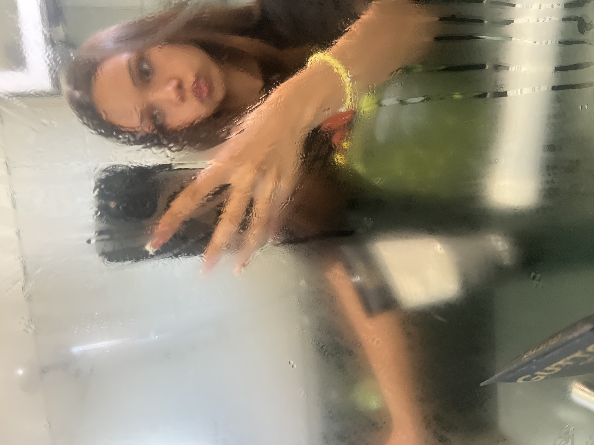 A young woman with long brown hair and a yellow bracelet is reflected in a fogged-up glass panel. She is holding a black smartphone with white fingernails. The environment appears to be a bus or train, judging by the blurry green and white horizontal lines and the angled "GUTS" lettering visible in the lower right corner. The location is unknown.
