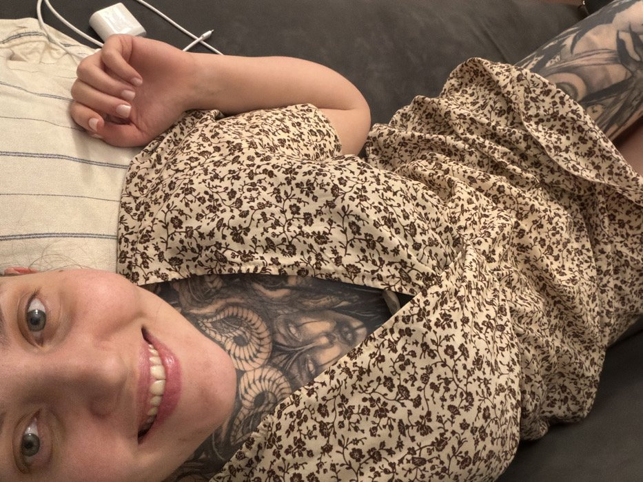 A woman lies on a couch, smiling. She wears a floral print dress and has extensive tattoos on her chest and leg. Her hand rests on a striped pillow. An electronic device and cord are near her hand. The background shows a dark-colored couch. Her head is positioned at the bottom left of the image, and her body extends upwards and to the right.