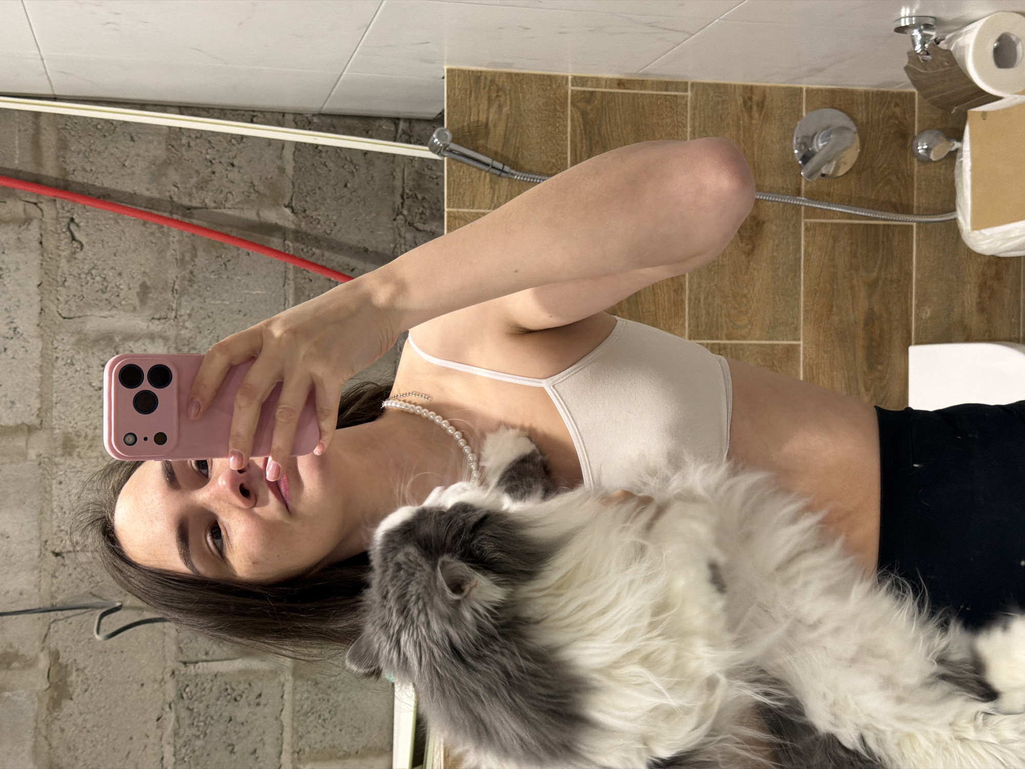 A woman is taking a selfie with a cat in a bathroom. The woman is wearing a white top, a pearl necklace, and black pants. She is holding a pink smartphone. The cat is gray and white and is nestled against her chest. The bathroom has tiled walls and includes a toilet, shower, and toilet paper dispenser. The image was taken in the city of Chaikovsky, Russia.