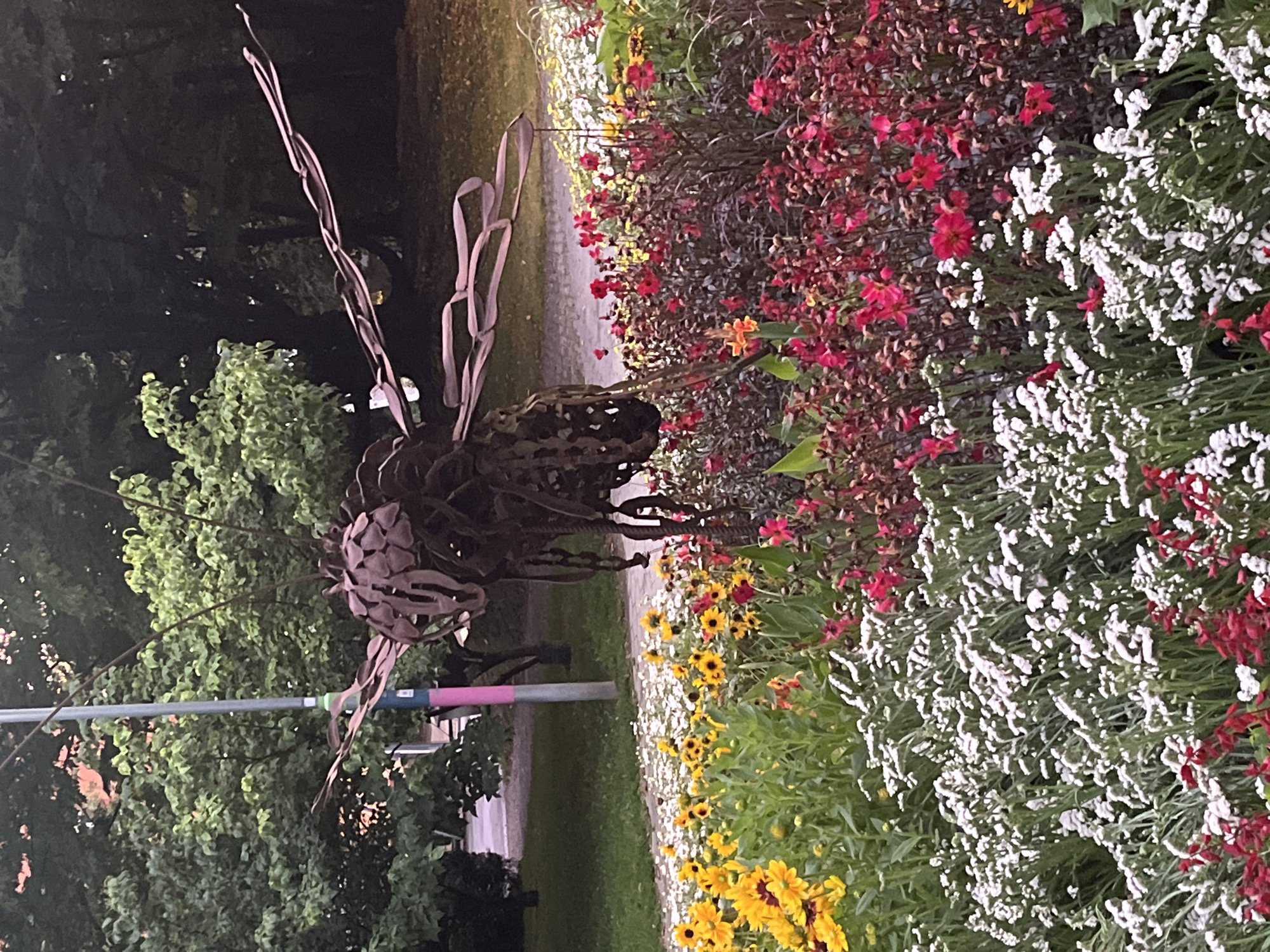 The image depicts a metal sculpture of an insect, possibly a bee or dragonfly, positioned in a flowerbed within a park. The sculpture is made of dark metal and has detailed wings and body. The flowerbed is filled with a variety of colorful flowers, including yellow sunflowers, red and pink flowers, and small white flowers. In the background, there are trees, a lamppost with colorful bands (blue, green, pink), and a paved path. The overall setting suggests an outdoor public space, likely a park or garden. The provided context states the location is in the city of Innsbruck, Austria. There are no people visible in the image.
