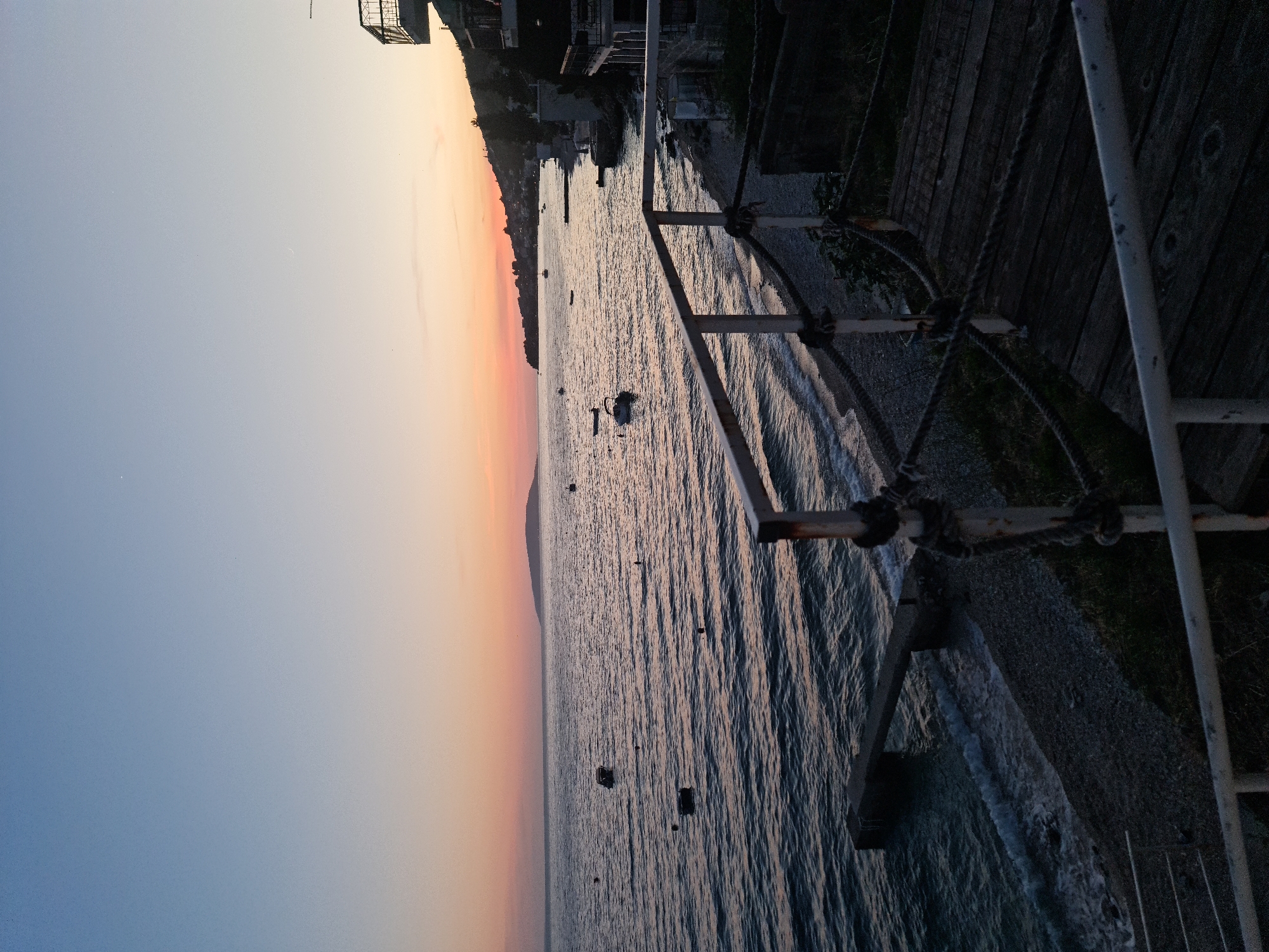 This is a landscape photograph taken at sunset in Brist, Croatia. The image captures a serene coastal scene with the sea in the foreground, gently lapping against a pebble beach. Several small boats are moored on the water, bobbing gently. A concrete pier extends into the sea from the left side of the frame.
In the mid-ground, the coastline of Brist is visible, dotted with buildings nestled into the hillside. The sky above is a gradient of soft colors, transitioning from a pale blue at the top to a warm orange and pink hue near the horizon, indicative of the setting sun. The light is soft and diffused.
The foreground features a wooden boardwalk and a white railing with thick ropes attached, suggesting a viewing platform or access point to the beach. The overall atmosphere is tranquil and picturesque.