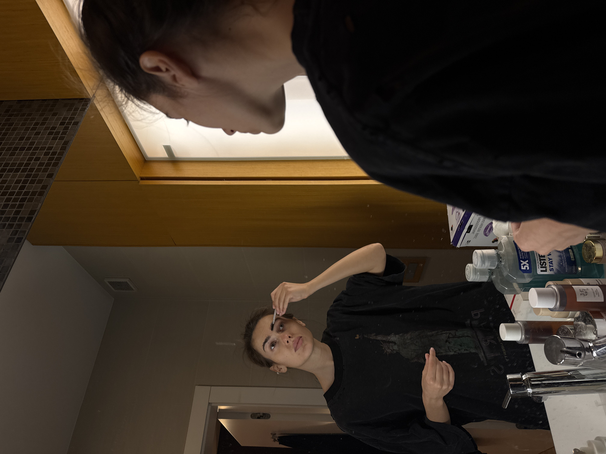 A young woman in a black t-shirt is standing in a bathroom, reflected in a mirror. She is holding a cotton swab near her eyebrow and appears to be grooming or applying something. Various toiletries, including bottles of mouthwash and skincare products, are arranged on the bathroom counter around the sink. The bathroom has tiled walls, a wooden cabinet, and a white door is visible in the background. The location is Unknown, Unknown.