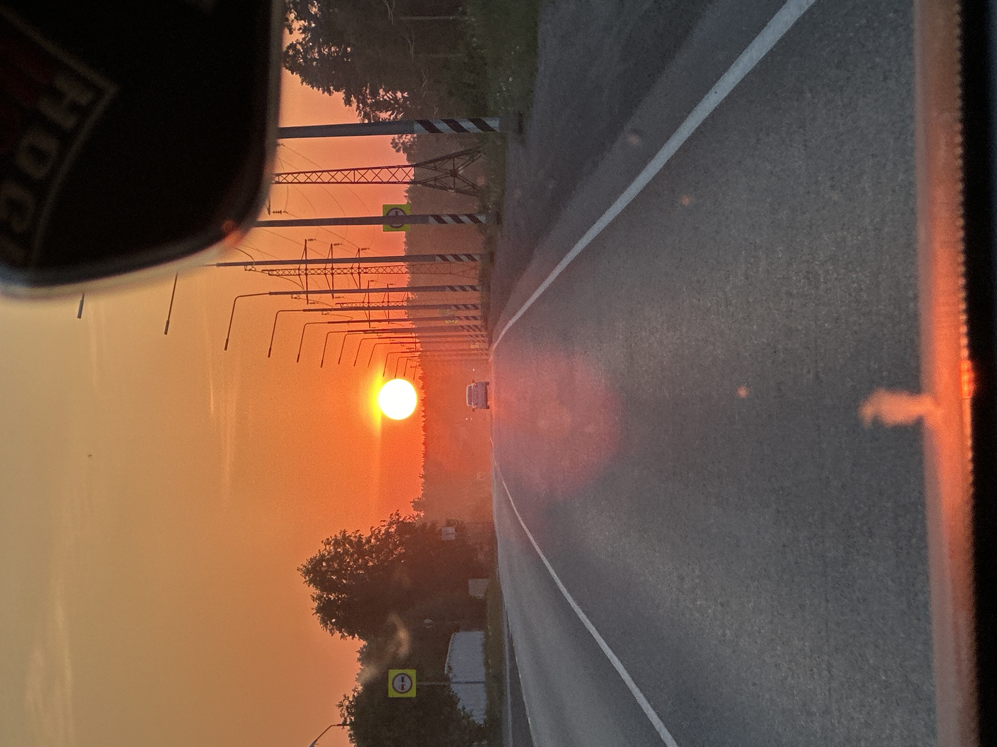 The image shows a road in Chaikovsky, Russia, at sunset. A car is visible in the distance. There are power lines and lampposts lining the road. A traffic sign is visible on the side of the road. The photograph appears to be taken from inside a vehicle.
