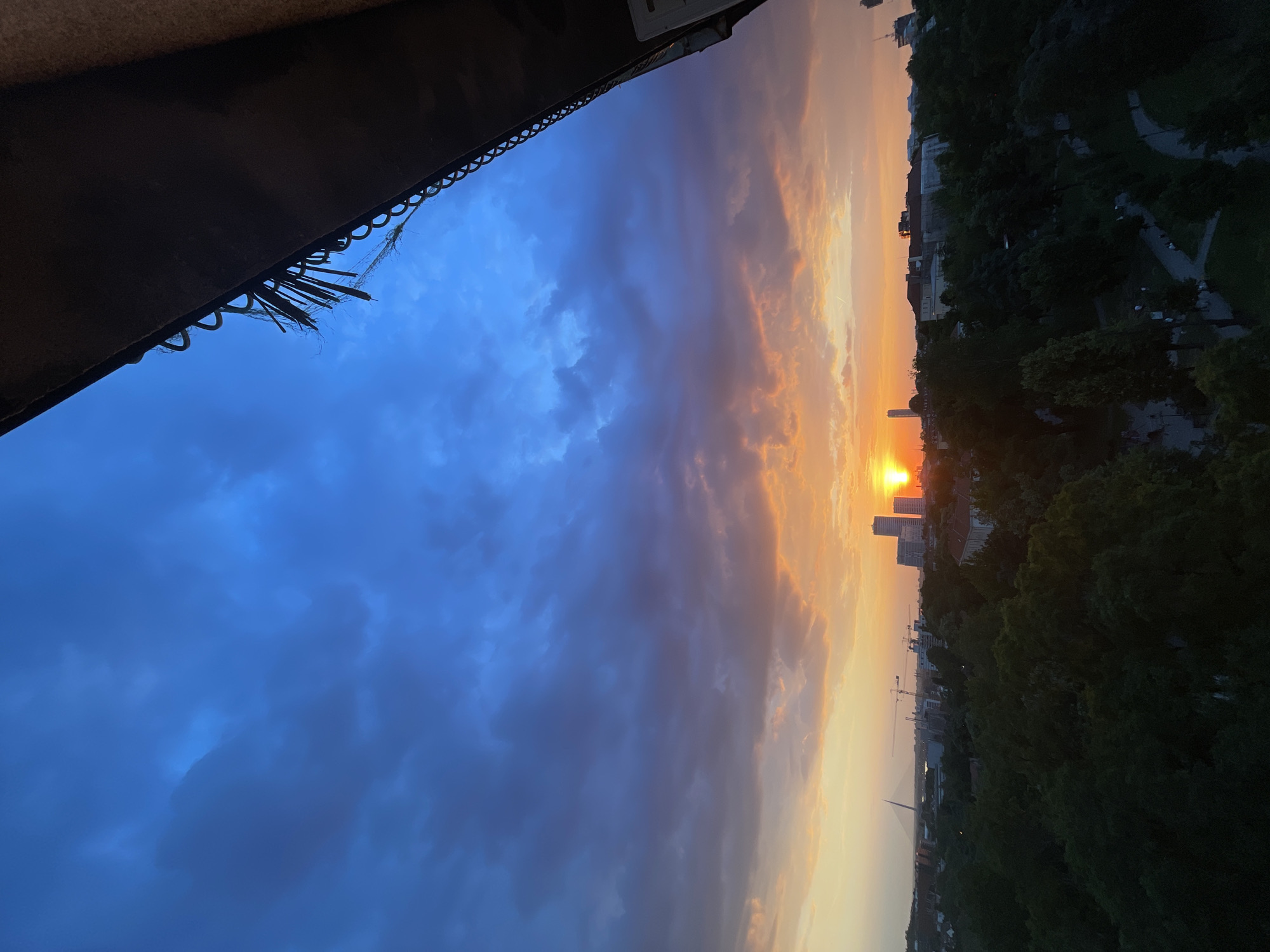 From an elevated perspective, possibly from a balcony, the image captures a sunset scene in Belgrade, likely from the Vracar Urban Municipality, Serbia. The lower portion of the frame shows a silhouette of a building’s edge. Above, the sky transitions from a deep blue on the left to orange hues near the setting sun, visible between buildings. Below the skyline, dense greenery suggests parkland. A distinct bridge is visible in the distance. A network of footpaths and roads run through the green areas.