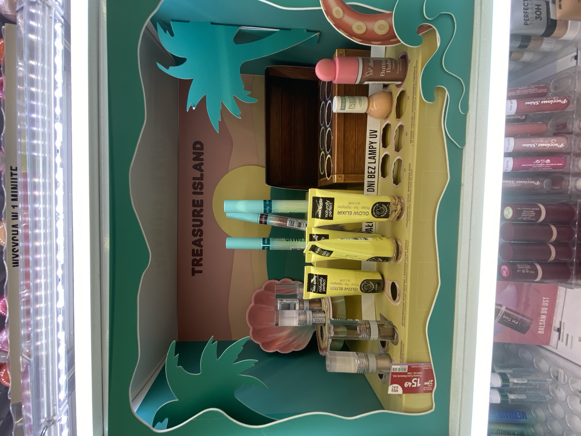 The image shows a retail display of makeup products. The display is themed as "Treasure Island" with palm trees and a treasure chest. Various makeup items are arranged on the display, including tubes of "Glow Elixir," bottles of mascara, lip glosses, and other cosmetic products. A price tag of 15.49 is visible on the lower left. The overall environment appears to be an indoor retail space, likely a cosmetics store. The lighting is bright, illuminating the products.