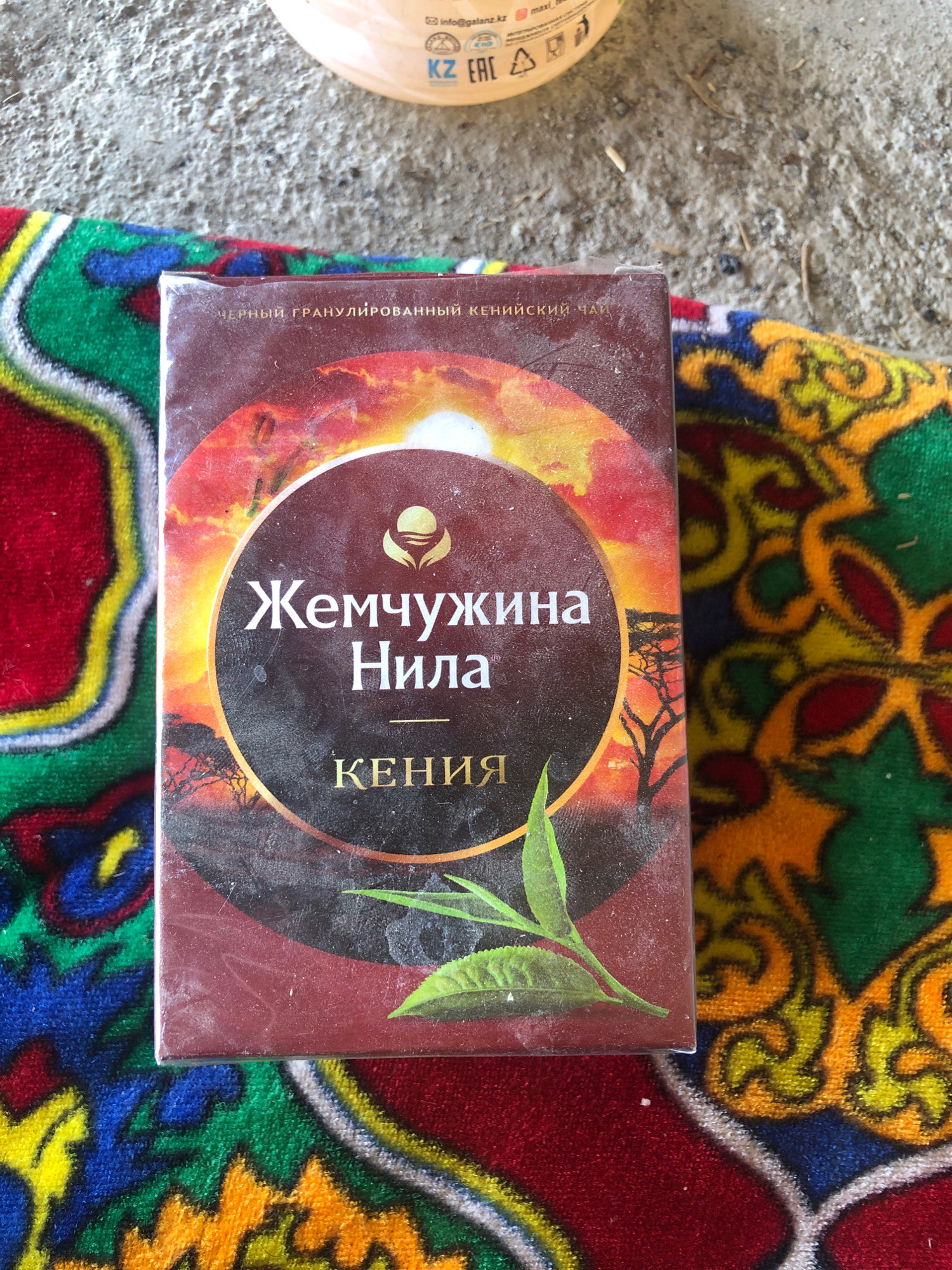 A close-up, slightly angled overhead shot captures a rectangular box of "Pearl of the Nile" black tea, predominantly brown and red with a circular emblem featuring a golden silhouette of a plant and sun. The text on the box is in Cyrillic script. The box is situated on a vibrantly colored woven blanket with geometric patterns in red, yellow, green, and blue, which rests on a textured, gray concrete surface. The lighting suggests an outdoor setting, possibly in Nookat, Kyrgyzstan, given the hints. The image has a slightly dusty or aged appearance, with visible smudges and dust particles on the tea box.