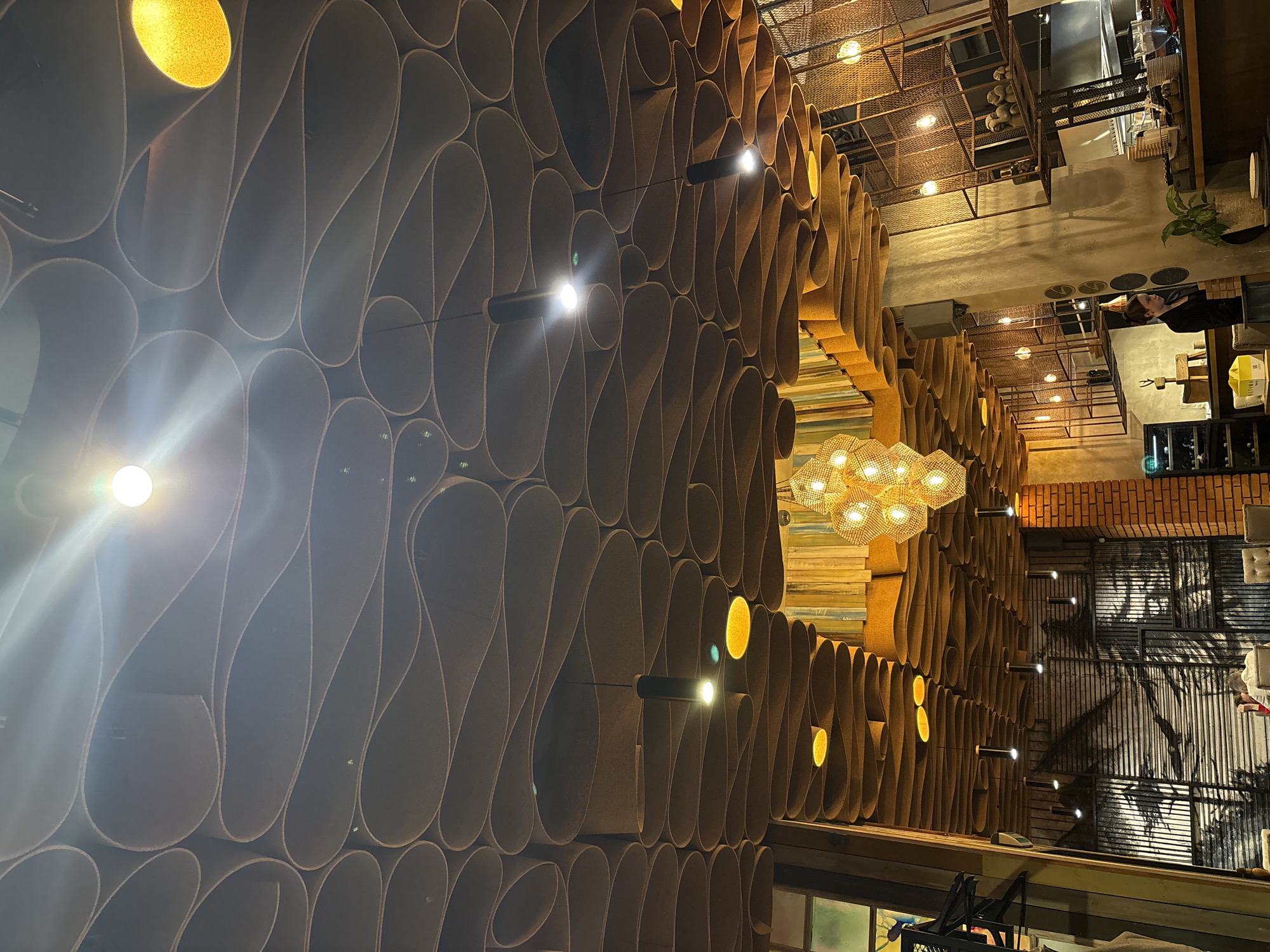 The image captures an interior view in Kazan, Russia, featuring a unique decorative wall made up of rolled-like structures that create a patterned design. The wall is illuminated by several light fixtures, including small cylindrical lights mounted directly on it and a large, decorative chandelier. The space appears to be a restaurant or bar, with glimpses of dining areas, kitchen equipment, and a bar counter visible in the background. On the right, a decorative painting or mural is visible behind a metal grill partition. There is a person visible behind the counter. Overall, the atmosphere suggests a modern and stylish establishment.