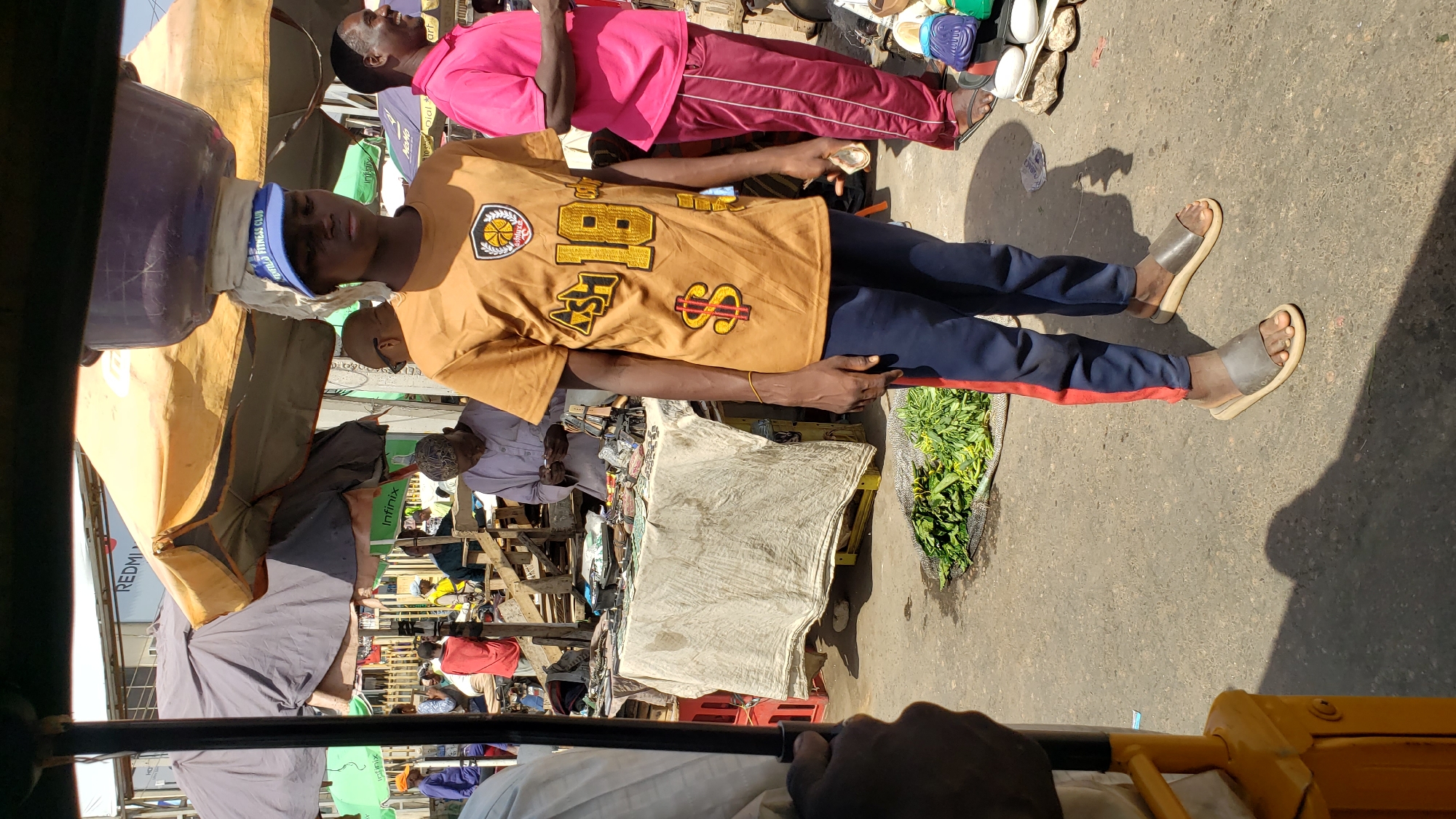 Captured from the perspective of a passenger inside a yellow motorized tricycle in what appears to be a market scene in Maiburiji, Nigeria. The foreground is partly obscured by the tricycle’s components and the passenger’s hand. The midground reveals a crowded marketplace with various stalls set up, displaying a wide range of goods. A man wearing a brown shirt with a basketball design, blue cap, and dark pants with a red stripe is standing casually beside one of the stalls. Another person in a pink outfit is positioned behind him. The stalls are covered with white cloths and assorted items, including shoes and vegetables. In the background, numerous people are moving around, browsing through the merchandise. Above the stalls, there are several sunshades providing cover. Overall, the scene conveys a lively and busy atmosphere typical of a market environment in Nigeria.