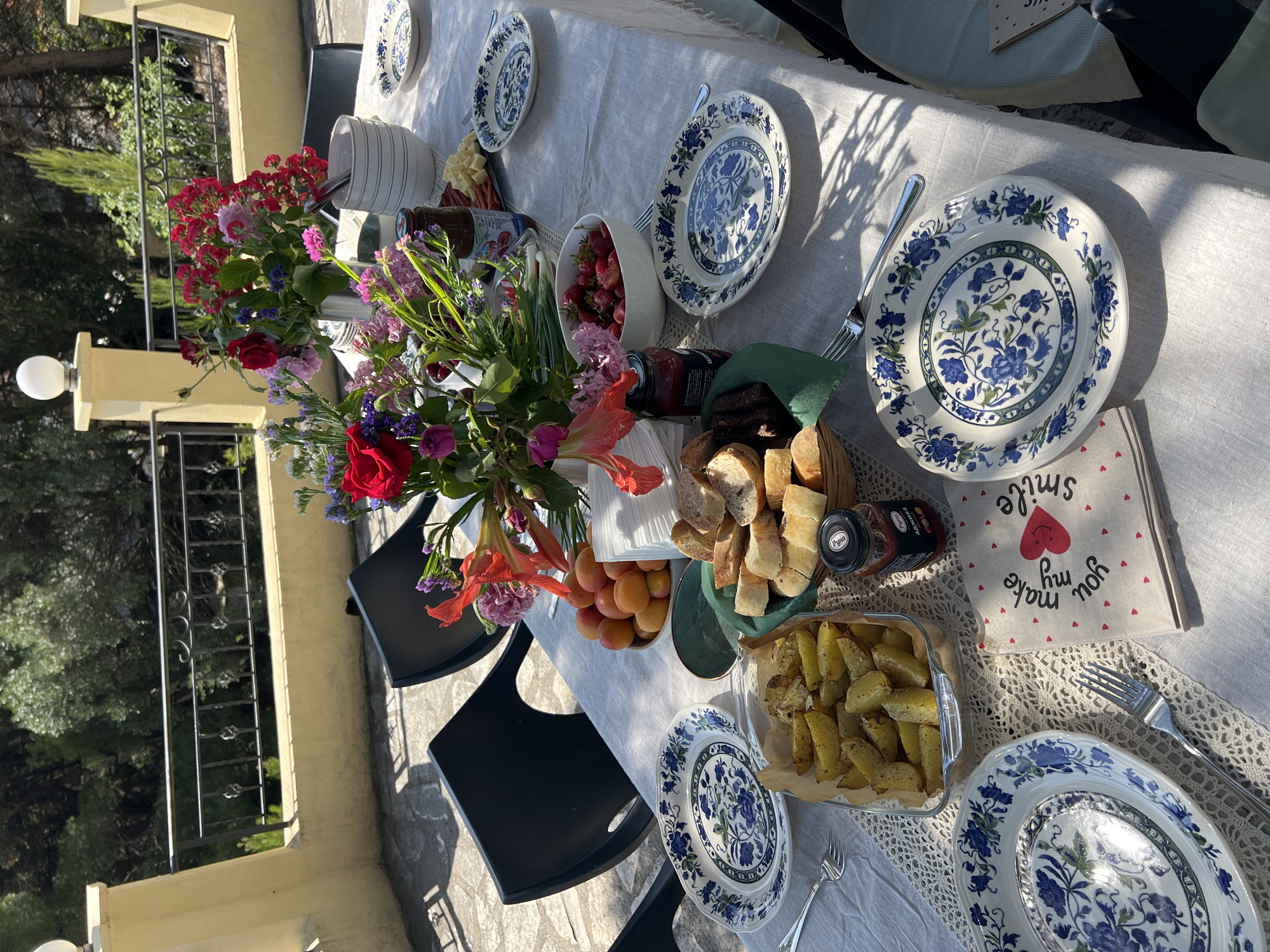 The image shows a set table on a balcony or terrace. The location is indicated to be Šušanj, Montenegro, although this cannot be confirmed visually. The table is covered with a light-colored tablecloth, possibly linen. It is set with several patterned blue and white plates and silverware. There is an assortment of food items, including bread, potatoes, strawberries, cheese, and various jams or preserves. Flowers in vases are placed along the center of the table. Several black chairs are arranged around the table. The lighting suggests a sunny day, with shadows cast across the scene. A balcony railing and foliage are visible in the background. There is a folded napkin with a "smile" and "you make my heart smile" message.