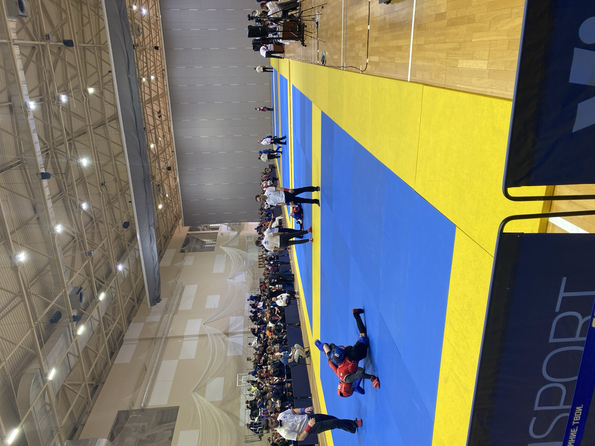 **Location:** Indoor sports arena in Sochi, Russia
**Scene:** A combat sports competition is taking place. A blue and yellow matted area dominates the scene, with two competitors engaged in a grappling match. One competitor is wearing red protective gear, while the other is wearing blue. A referee is positioned nearby, observing the action.
**People:** In addition to the competitors and referee, a crowd of spectators is seated along the sidelines, watching the competition. There are additional officials and staff present, walking along the perimeter of the matted area. A media setup with cameras is visible in the background.
**Objects:** The arena features a high ceiling with exposed beams and lighting fixtures. Banners and signage are present, including one with the word "SPORT" partially visible in the foreground.