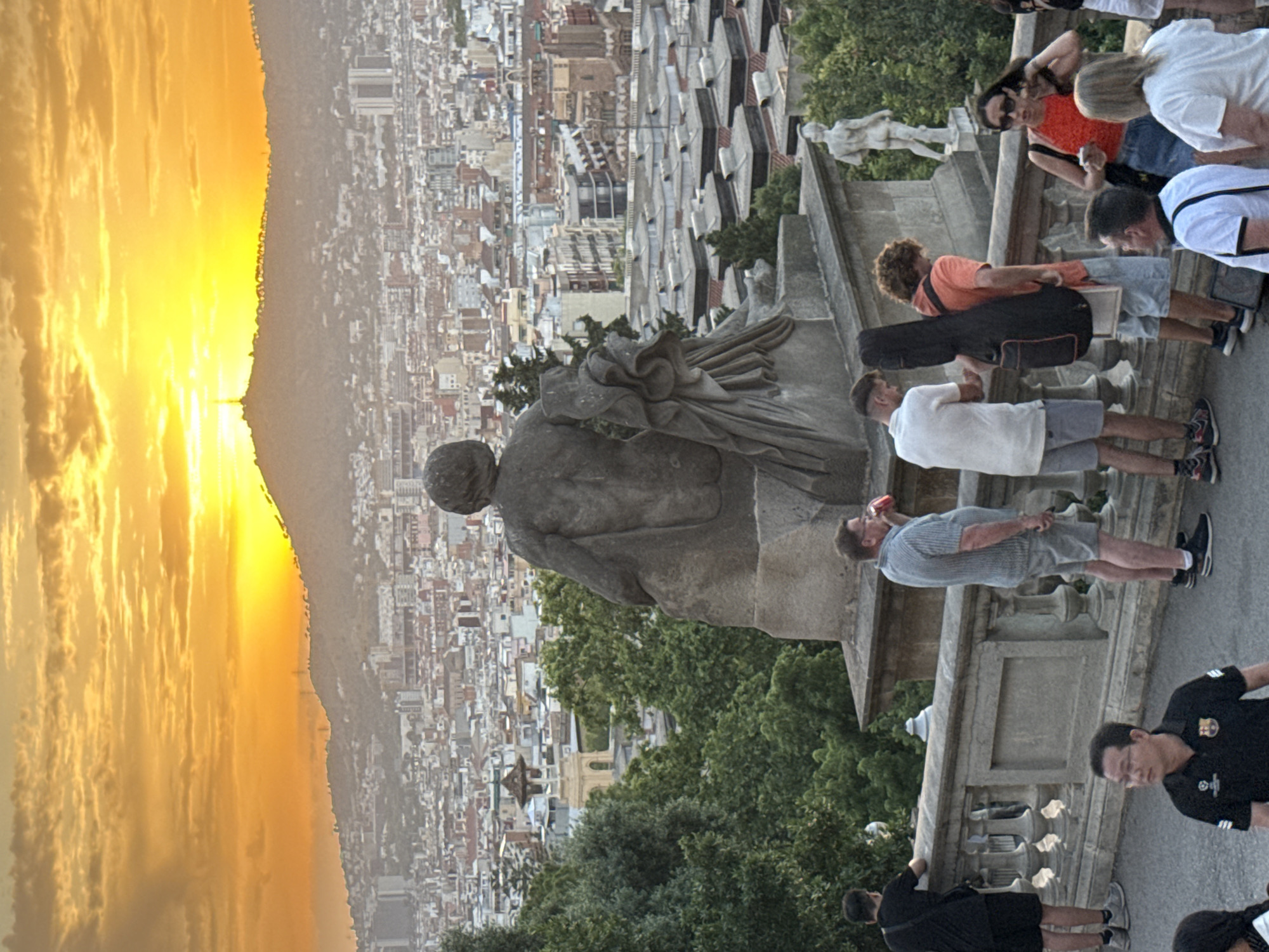 The image depicts a scenic overlook in Barcelona, Spain, at sunset. In the foreground, a large stone sculpture of a seated figure faces away from the viewer, looking towards a sprawling cityscape. Several people are gathered along a stone railing in front of the sculpture. From left to right, a man in a black t-shirt and shorts stands facing the railing. Next to him, a man in a grey striped shirt and shorts drinks from a red cup. Further right, a man in a white t-shirt and shorts stands with a guitar case slung over his shoulder. He is standing next to a young man in an orange t-shirt and light blue shorts, also with a guitar case. A woman in a red top and denim skirt stands to the right, holding a phone. Behind the railing, on a lower level, a man in a black polo shirt with a logo stands looking forward. Another person, only partially visible, is in the bottom left corner. The background reveals the extensive urban landscape of Barcelona, with buildings stretching towards a distant hill topped by a structure, silhouetted against a vibrant orange and yellow sunset sky. Lush green trees frame the view from the overlook.