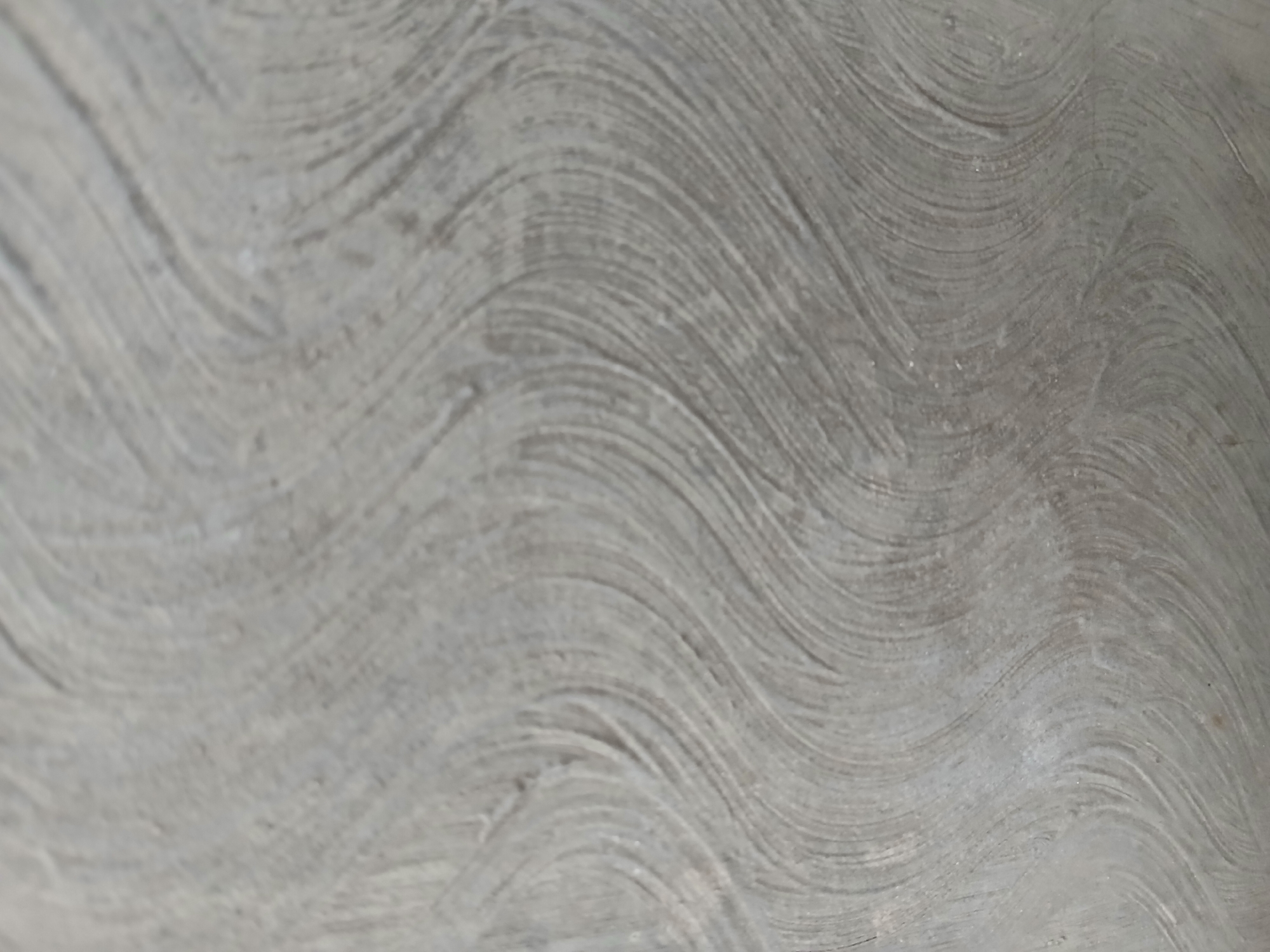 The image displays a close-up, abstract view of a textured surface. The surface is predominantly gray with visible swirling patterns that suggest a brushed or troweled finish, possibly concrete or plaster. The lighting casts subtle shadows that accentuate the undulating texture. There are no people, identifiable objects, or discernible actions present. The context of Tenggarang, Indonesia is not visually represented in this abstract image.