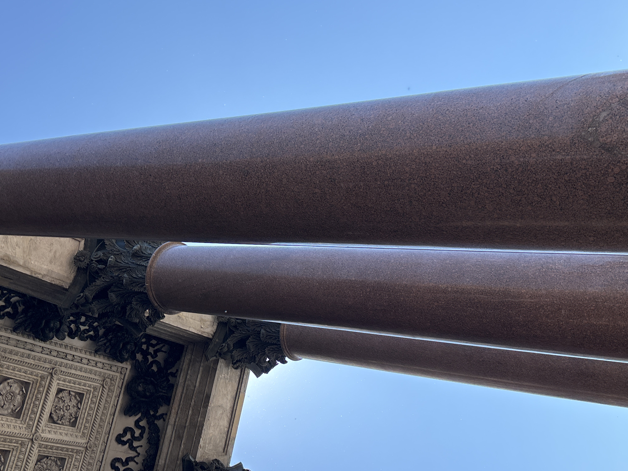 This is a low-angle, close-up, outdoor photograph of massive, polished red granite columns supporting an ornate architectural element, likely the portico of a grand building. The columns are thick and cylindrical, with subtle vertical lines indicating their polished surface. The architectural detail above is intricate, featuring dark, sculpted ornamentation resembling acanthus leaves and cherubic faces, set against a textured ceiling with square recessed panels. The bright, clear blue sky fills the background, with sunlight creating a slight lens flare in the upper left quadrant. The image appears to capture a detail of a significant historical structure. Given the monumental scale of the columns and the classical architectural style, this scene is likely located at **Saint Isaac's Cathedral** in Saint Petersburg, Russia, which is famous for its large red granite columns and richly decorated interior. There are no people visible in the image.