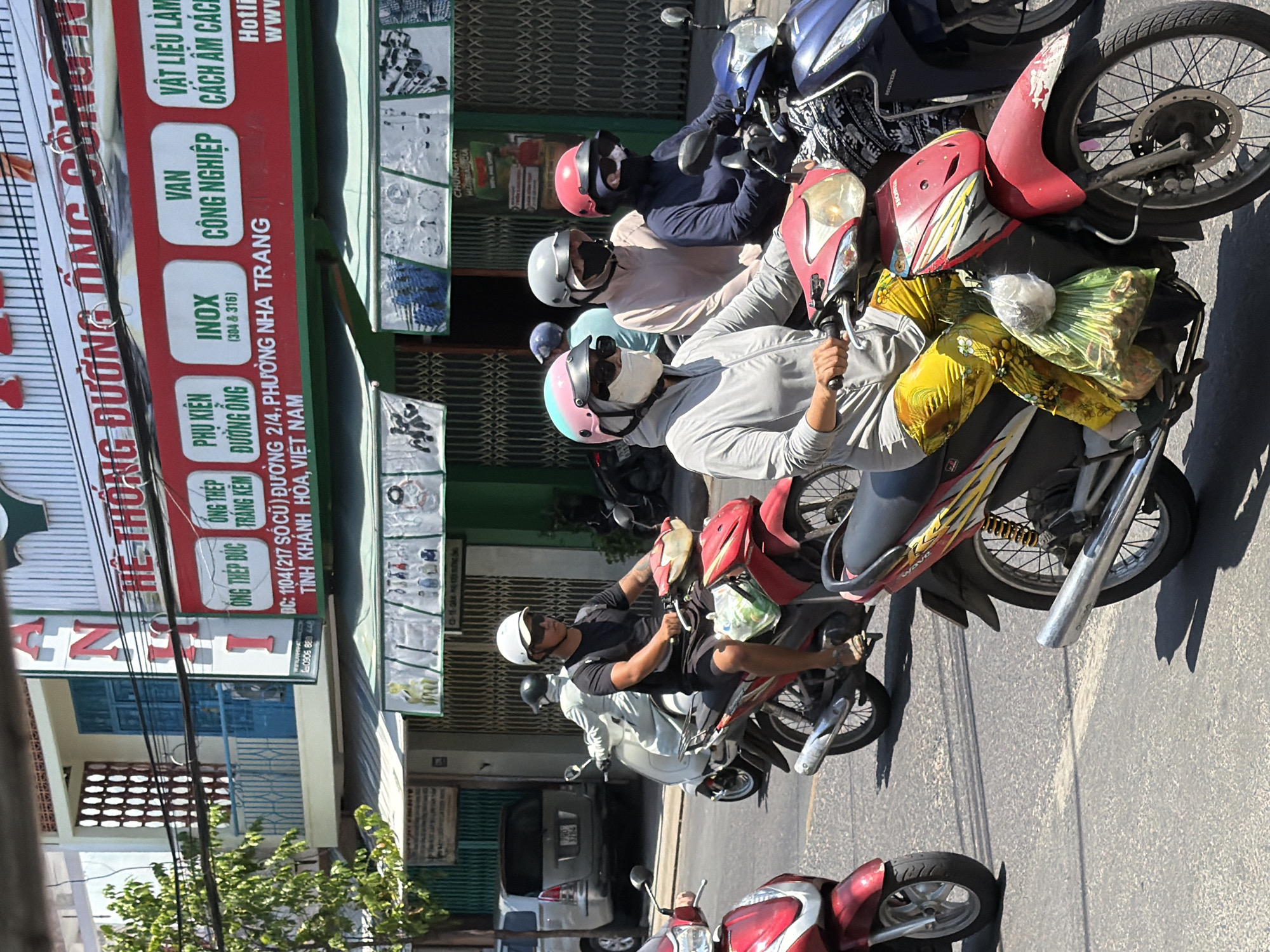 A group of people on motorcycles are riding down a street in Nha Trang, Vietnam. The motorcycles are a mix of red and blue, and the riders are wearing helmets and masks to protect themselves from the sun and dust. The street is lined with buildings, some of which have awnings and signs in Vietnamese. The buildings appear to be shops and businesses, and there is a lot of activity on the street. One of the signs on the building reads "ANH", indicating a business name. The overall scene is one of a busy urban environment in Vietnam. The image captures a snapshot of daily life in Nha Trang, with people commuting and going about their day on their motorcycles.