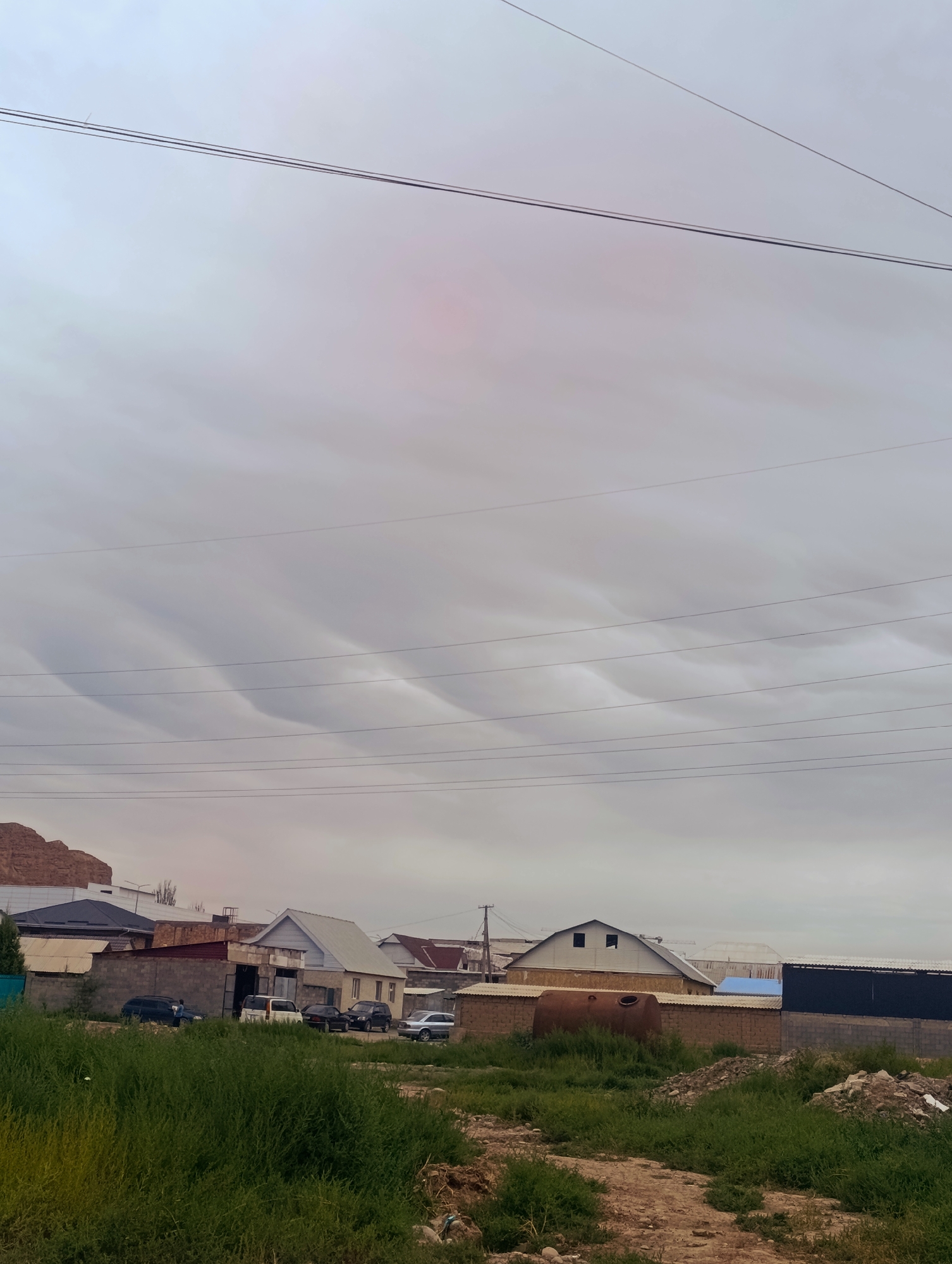 The image shows a residential area in Naryn City, Kyrgyzstan. The sky is overcast with distinctive wave-like cloud formations. Several power lines are visible crisscrossing the upper portion of the frame. In the mid-ground, a cluster of houses and buildings are situated, some appearing to be under construction. Several cars are parked in front of the houses. The foreground consists of unkempt vegetation and dirt. The overall scene depicts a somewhat rural or developing urban environment under a dramatic sky.