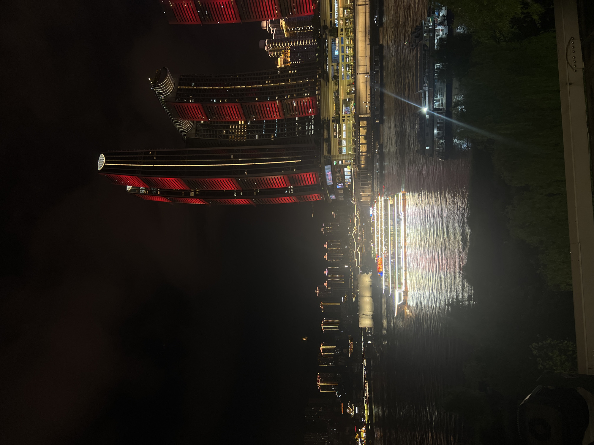 This is a nighttime photograph of a riverfront cityscape in Jiangbeicheng Subdistrict, Chongqing, China. The image captures a wide river with several boats illuminated by artificial lights. In the background, prominent skyscrapers are lit up with red and yellow lights, creating a vibrant urban spectacle against the dark sky. One of the most striking features is a group of modern, tall buildings with distinctive architectural designs, prominently displaying red LED lighting. A large passenger boat, also brightly lit, cruises on the river, its lights reflecting on the water. Other smaller boats are also visible. The foreground shows a dark area with some vegetation, possibly trees, and a railing. The overall atmosphere is one of a lively, modern city at night.