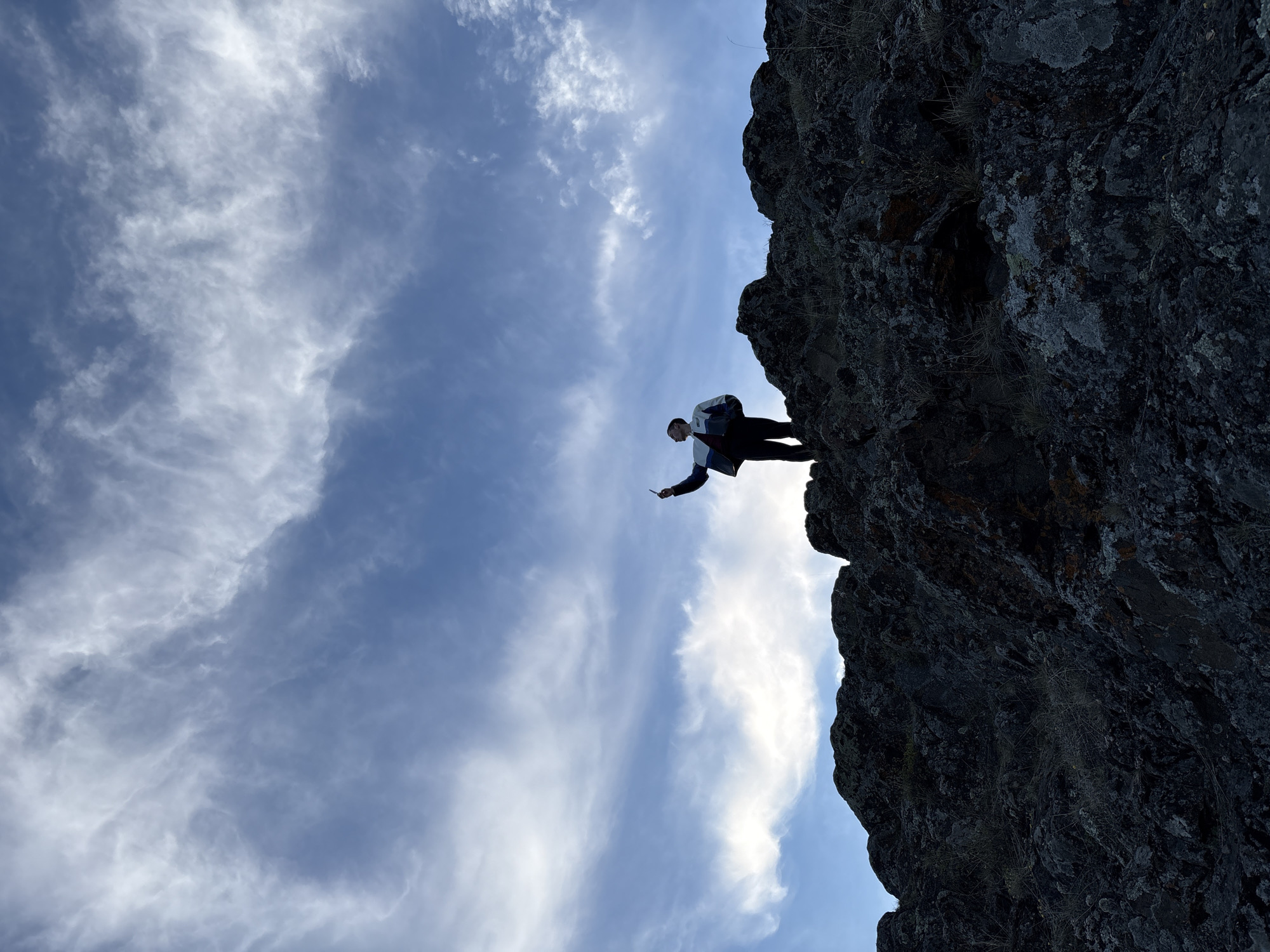 A low-angle shot reveals a person standing on the edge of a dark, rocky cliff against a backdrop of a partly cloudy blue sky. The person is wearing a light-colored jacket over a darker shirt, dark pants, and is holding something in their outstretched hand. The location is speculated to be near the city of Невон, Russia.