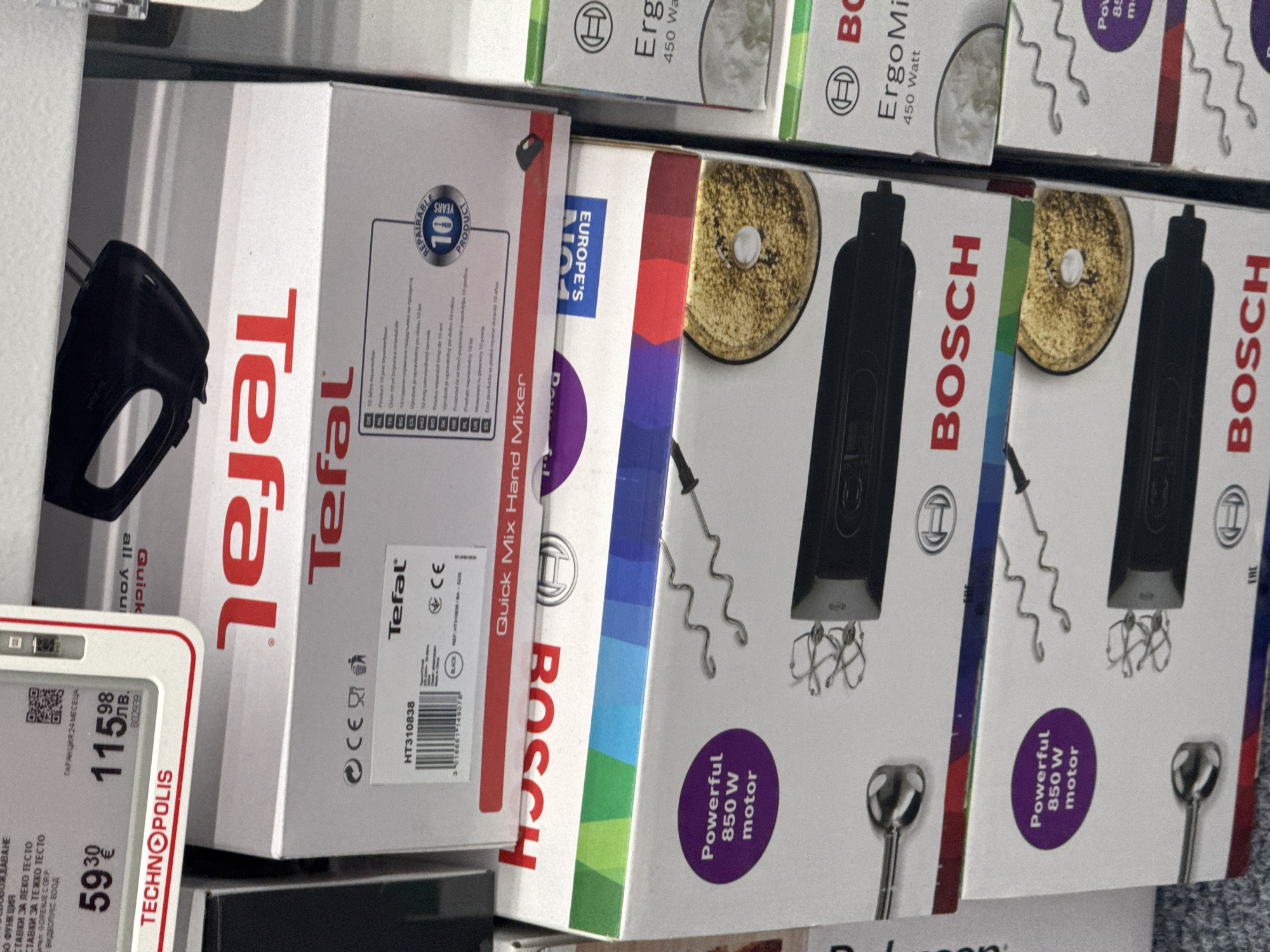 The image shows a shelf in a retail store, likely in Lovech, Bulgaria, based on the "TECHNOPOLIS" sign and Bulgarian text on the price tag. The shelf is stocked with hand mixers. Featured prominently are boxes of "Tefal" and "BOSCH" hand mixers. The Tefal box is primarily white and red with the brand name displayed vertically. The Bosch boxes are predominantly white with red and blue accents, also displaying the brand name prominently. The Bosch mixers are advertised as having a "Powerful 850 W motor." A price tag shows a product for "59.30" and "115.98." The environment is a typical retail setting, with multiple units of the same appliances stacked on the shelf.