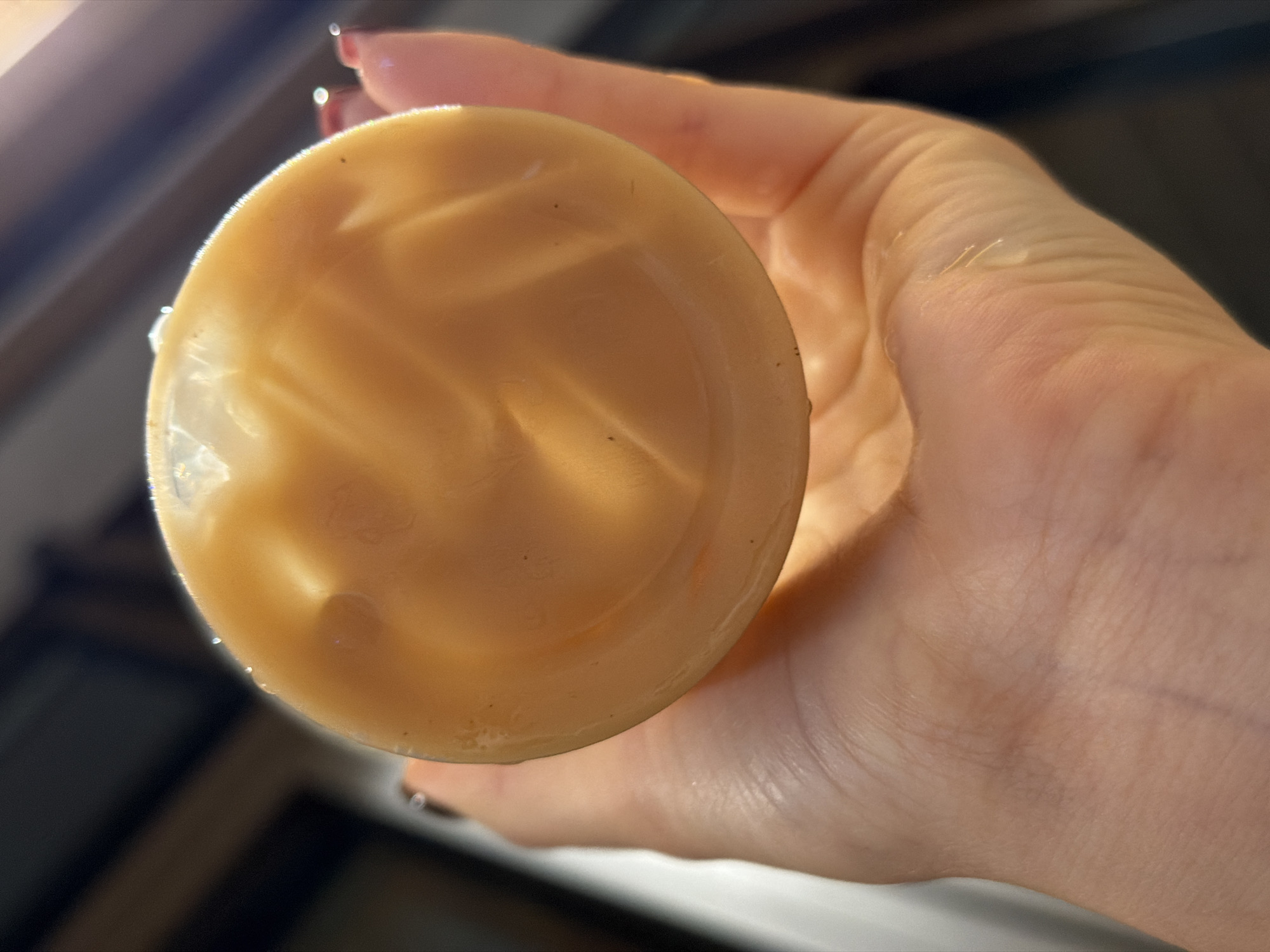The image shows a close-up, overhead shot of a person's hand holding a round, yellow object. The object appears to be a container or lid with a slightly textured surface, possibly a jar or a bottle cap. Swirls and indentations are visible on the surface of the yellow object, suggesting it might be made of plastic or a similar material. The hand holding it has fair skin and appears to be illuminated by warm light, casting subtle shadows. The fingers are slightly curved around the object. There are hints of red nail polish on one of the fingernails. The background is blurred and out of focus, showing a dark, indistinct area that could be an interior or a dimly lit outdoor environment. There are some faint reflections and light sources visible in the background. The focus is entirely on the hand and the yellow object.