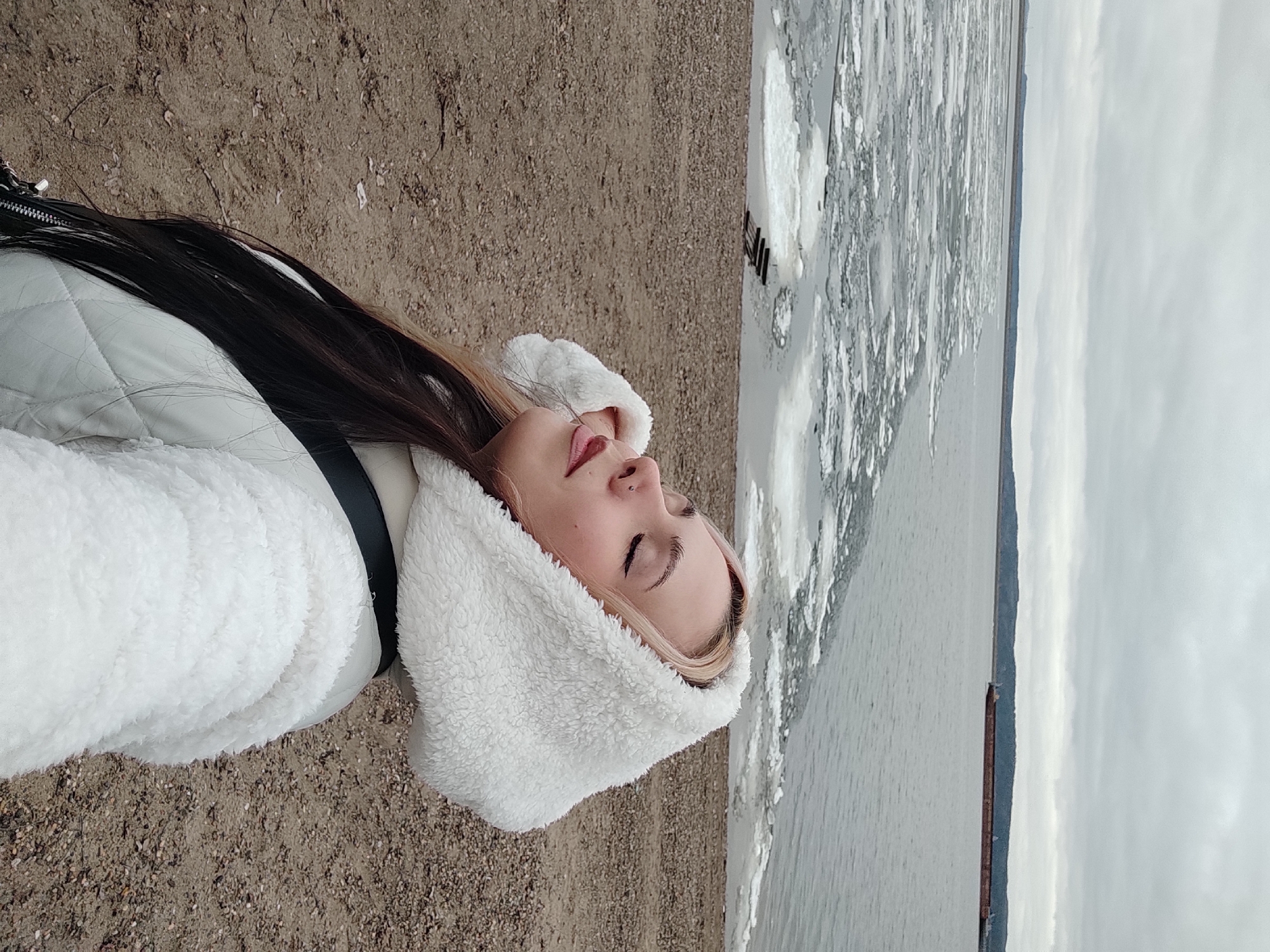 The image shows a woman lying on a beach in what is likely the city of Chaikovsky, Russia. She is wearing a white jacket and a furry white hood. She has long dark hair with blonde highlights and is lying on her back with her eyes closed. The beach is sandy and next to a body of water covered with ice floes. The sky is overcast. The location is likely the Kama River in Chaikovsky, Russia, given the presence of the body of water and the evidence of ice formation.