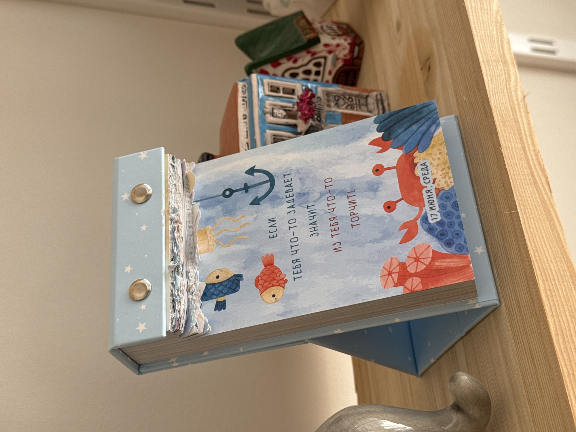 The image shows a blue desk calendar with a nautical theme. The calendar is open to a page with illustrations of fish, jellyfish, an anchor, coral, and a crab. There is text on the page written in Russian, which translates to "If something is bothering you, it means something is stuck!" Below the illustration, the date "17 ИЮНЯ, СРЕДА" (June 17, Wednesday) is visible. The calendar is placed on a wooden shelf. In the background, there are two small decorative houses, one blue and white, and another green and white, resembling model buildings. To the left of the calendar, a portion of a light-colored ceramic object, possibly a sculpture or figurine, is visible. The lighting suggests the scene is indoors, with natural light coming from the right. There are no people or animals actively engaged in the scene. The overall context suggests a domestic or personal space, likely a home or office. There is no information to place this scene specifically in the city of Izhevsk, Russia, or any other specific city.