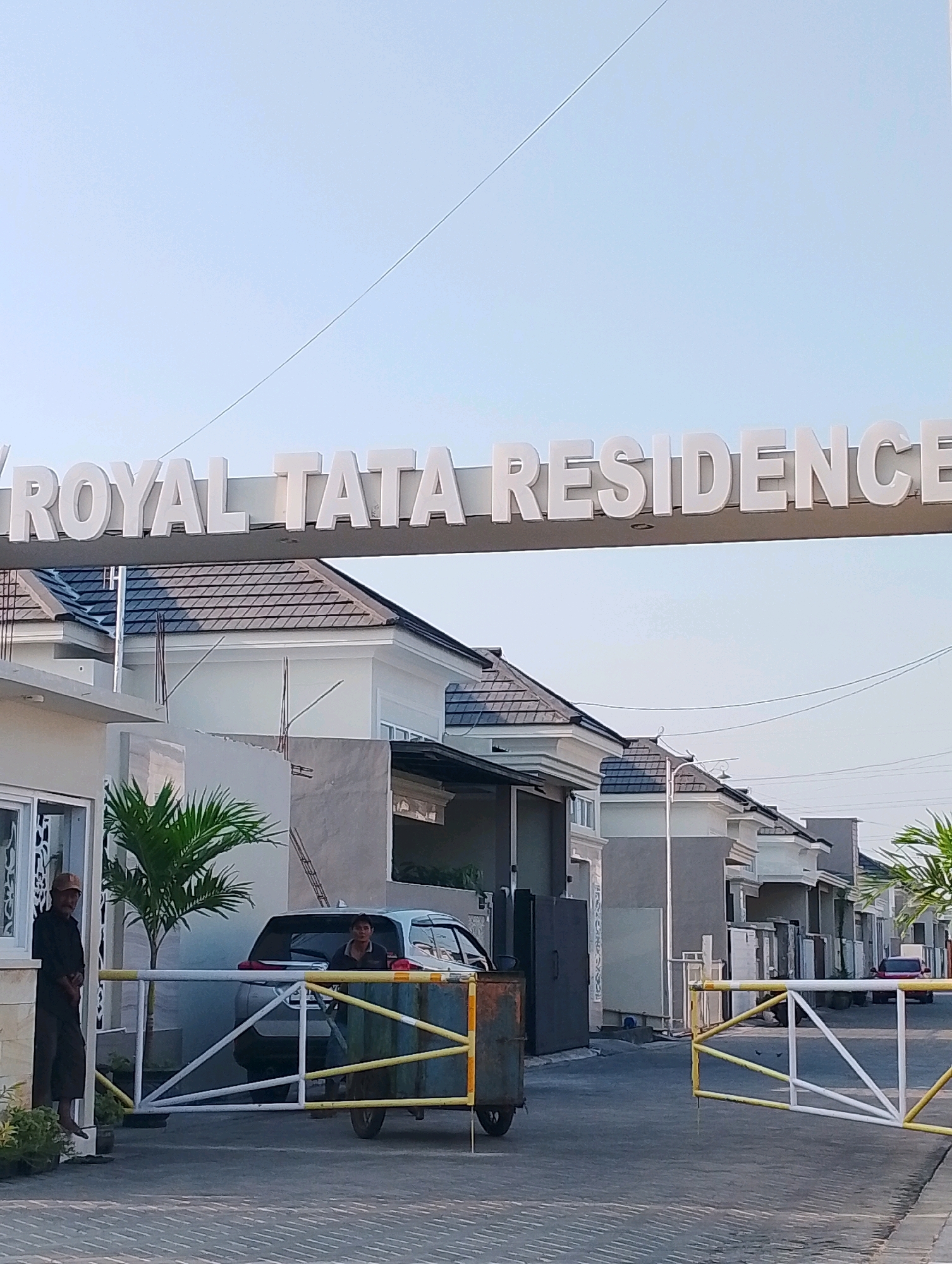 An exterior, medium-shot photograph captures the entrance of the "ROYAL TATA RESIDENCE" in Probolinggo, Indonesia, under a clear sky. The sign, made of white, raised lettering, is mounted on a horizontal structure above the entrance.
The entrance is a paved road leading into a residential area. A metal barrier gate, painted white and yellow, is partially open, allowing passage. A security guard, dressed in a dark shirt and pants with a cap, stands to the left of the gate, near a small structure that appears to be a guard post. Another individual, likely a security guard as well, is visible behind the gate, partially obscured by a vehicle.
A silver SUV is parked behind the barrier gate. In the background, modern houses with tiled roofs are visible, lining the street. A small palm tree stands to the left of the entrance, near the security guard. The overall scene suggests a secure and well-maintained residential community entrance.
