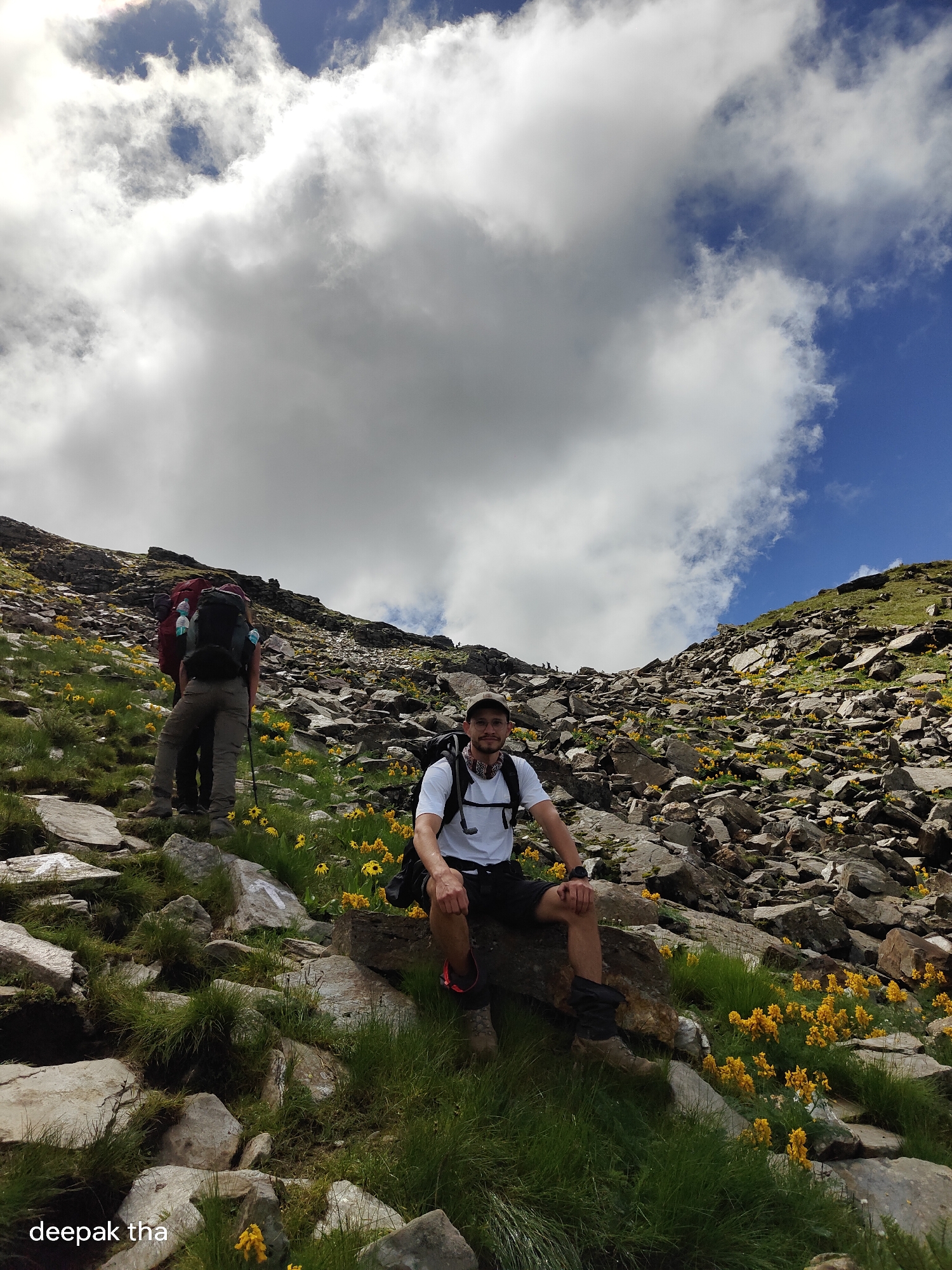 A low-angle, outdoor, full-shot photograph captures a mountainous landscape with two hikers. The sky is filled with dramatic clouds, with patches of blue visible. The terrain is rocky and steep, covered with green grass and scattered yellow wildflowers.
In the foreground, a man wearing a white t-shirt, black shorts, and hiking boots is seated on a large rock. He is looking towards the camera and smiling. He has a backpack on and a bandana around his neck. His legs are bare, and he is wearing gaiters on his lower legs.
Further up the rocky slope, another hiker is seen from behind. This person is wearing a red backpack and brown pants, and they are holding a hiking pole.
The location appears to be in the mountains near Bashisht, India, given the presence of yellow wildflowers commonly found in Himalayan regions. The image conveys a sense of adventure and the beauty of nature.