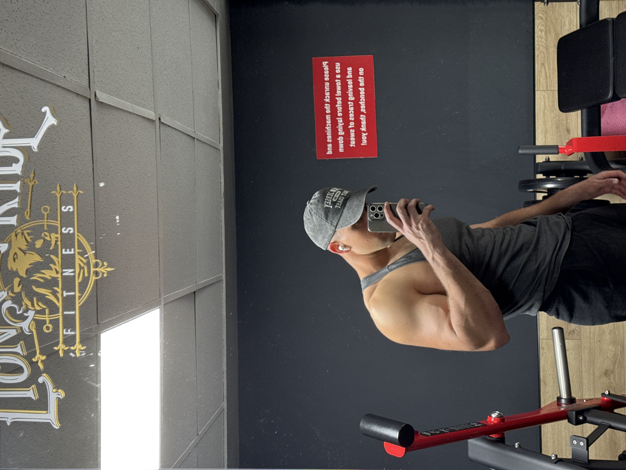 A man is taking a mirror selfie inside a gym. The image is rotated 90 degrees clockwise, showing the scene sideways.
The subject is a muscular man, wearing a grey tank top, black shorts, a grey baseball cap, and white wireless earbuds. He is holding a smartphone, capturing his reflection.
The setting is an indoor gym, featuring dark grey or black walls, one of which is a large reflective surface (likely a mirror or glass panel). The floor is a mix of dark and light wood-patterned sections. Gym equipment, predominantly red and black, is visible in the reflection, suggesting a leg press or similar resistance machine.
Notable details include the man's visible arm musculature and the reflection creating a sense of depth. The general lighting suggests it is likely daytime.
Visible text includes a gold and white logo on the reflective wall that reads "LONG HARDY FITNESS" (with a lion's head design). A red rectangular sign, also reflected in the mirror, displays mirrored text that, when corrected, reads "Please unlock the machine" and "Use a towel before leaving." The remaining text on the sign is less clear but appears to be further gym etiquette rules.