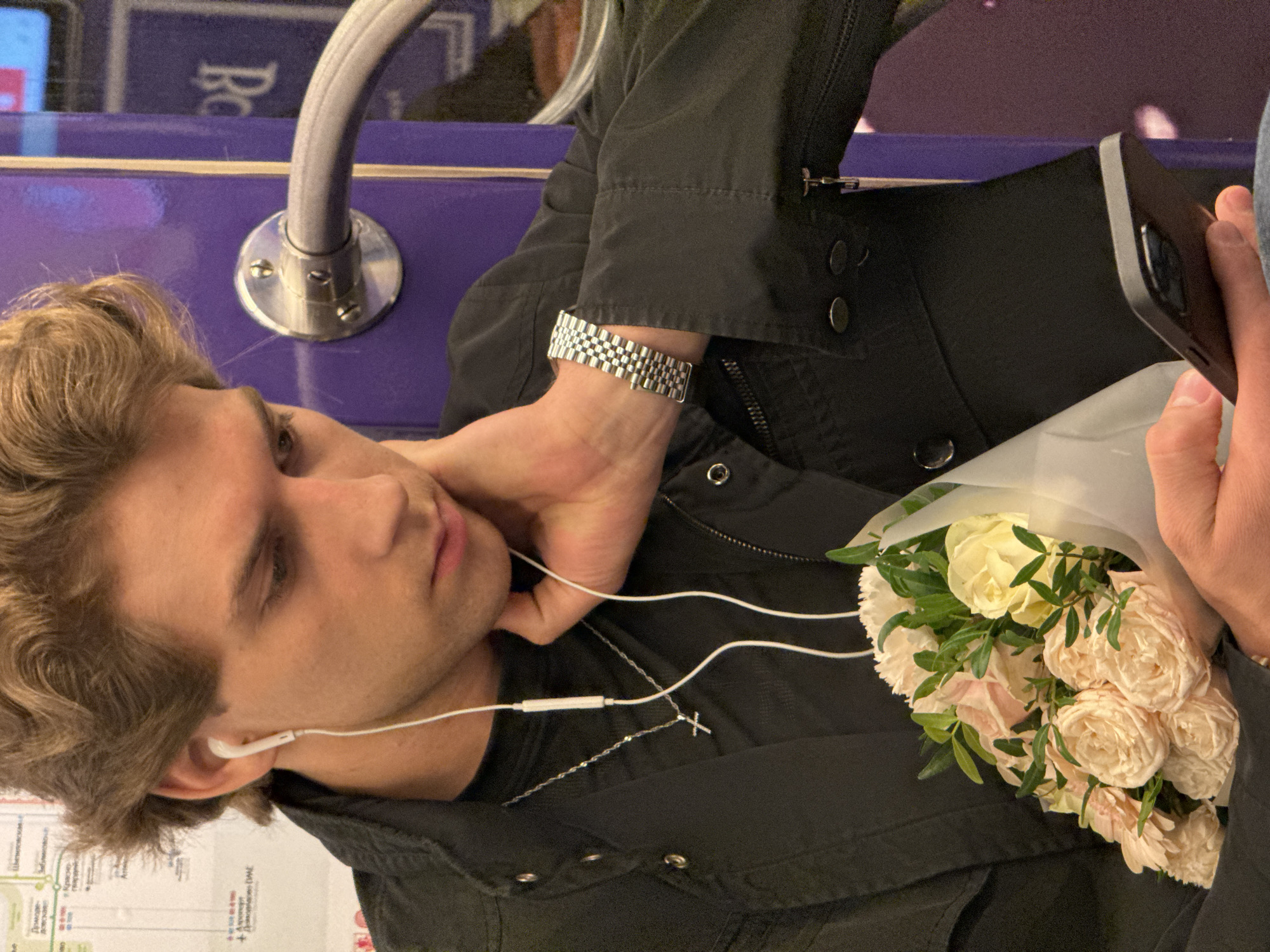 A young man sits on public transport, possibly a train or bus, holding a bouquet of light-colored roses. He is wearing a dark jacket, a metal watch on his left wrist, and a silver chain with a cross pendant. He has earbuds in and is looking down at a phone he holds in his right hand. The transport's interior includes a purple panel with a silver grab bar. The Russian writing visible suggests this is in Moscow, Russia. A map of the Moscow Metro is partially visible in the bottom left corner.