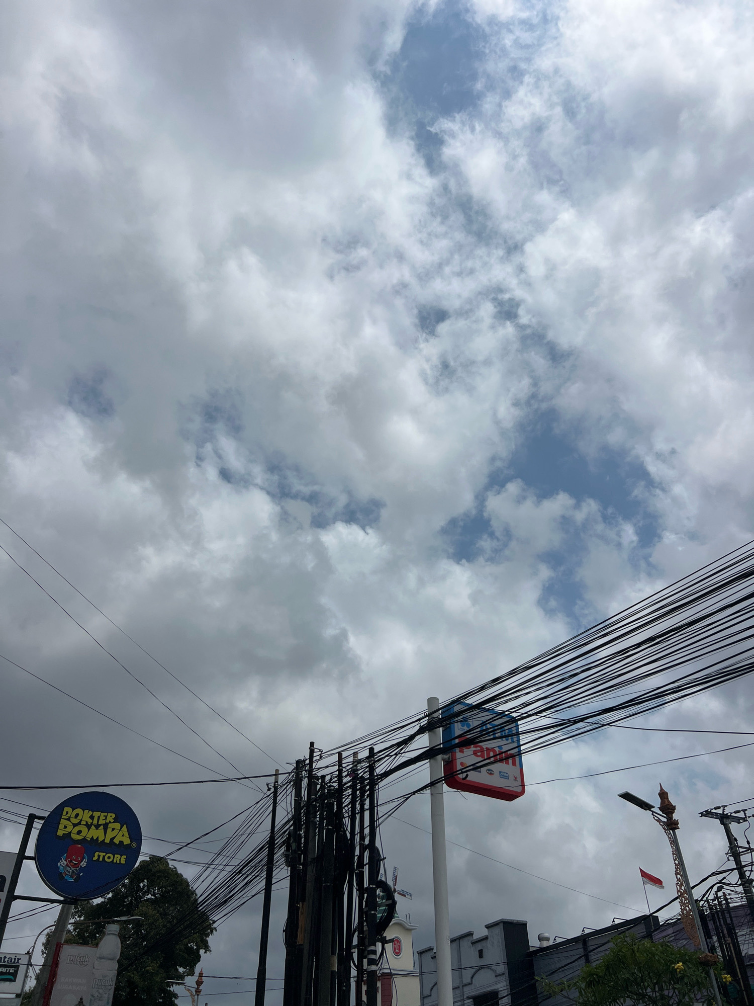 This is a low-angle, full outdoor shot of a cloudy sky over a city street in Denpasar, Indonesia. The sky is filled with a mix of white and gray clouds, with patches of blue sky visible. A dense network of black electrical wires crisscrosses the frame, supported by utility poles.
On the left side of the image, a circular blue sign with yellow lettering reads "DOKTER POMPA STORE" and features a cartoon character. To the right of this sign, a rectangular red and white sign is mounted on a white pole, with some text and logos partially visible. In the background, several buildings are visible, including one with a white steeple and a windmill-like structure, and another with a gray facade. A street light with an ornate base is also present, and a small Indonesian flag is flying. Lush green trees are seen on the left and right edges of the frame. The overall impression is that of a typical urban street scene with a focus on the sky and overhead infrastructure. There are no people or animals visible in the image.