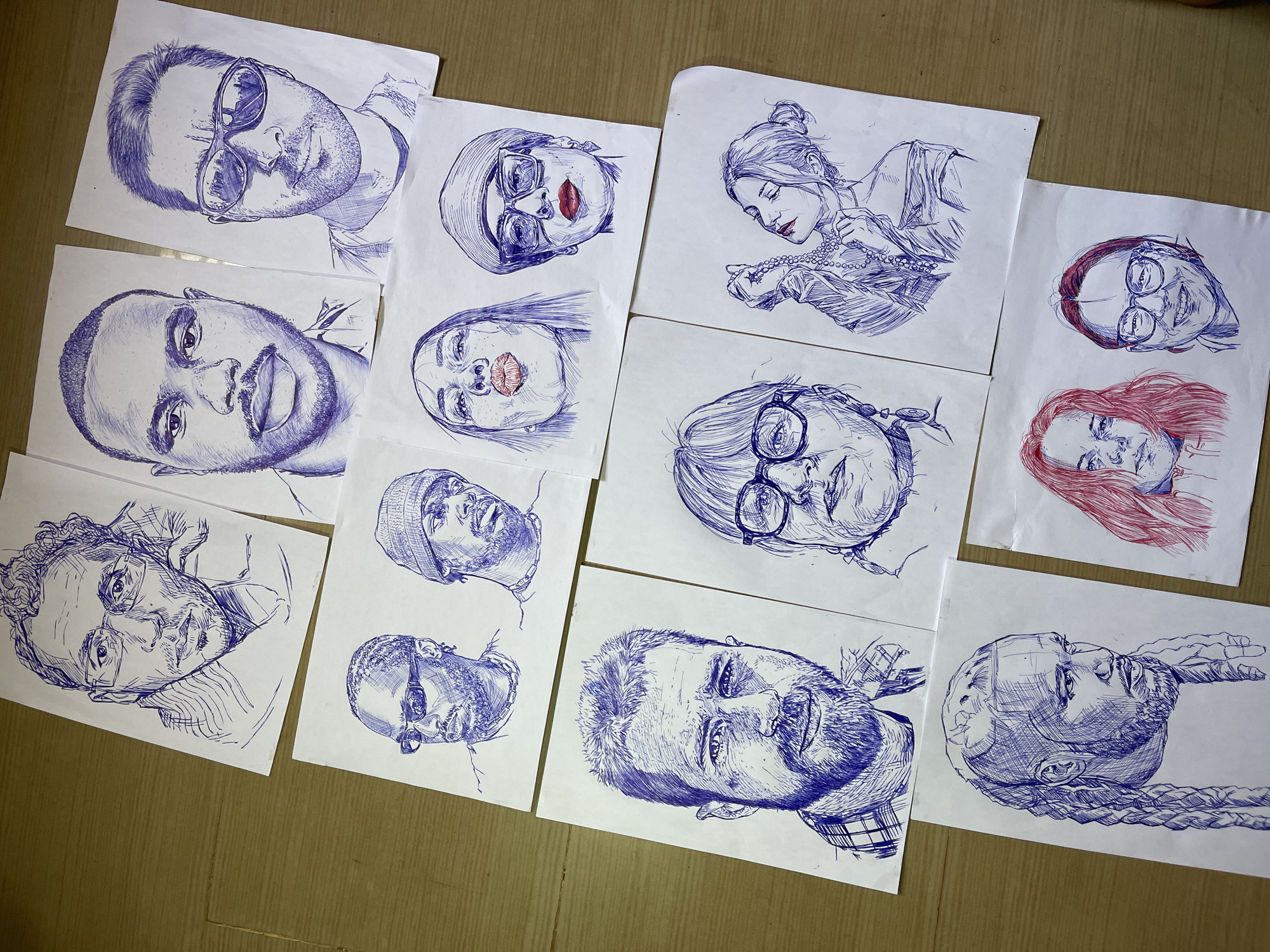 The image displays a collection of pen-and-ink drawings of people, arranged on a wooden surface that appears to be a floor. The drawings are primarily portraits, featuring a variety of individuals with different expressions and hairstyles. Some of the subjects wear glasses, while others are depicted with hats or head coverings. One drawing shows a woman adorned with a pearl necklace, and another features a person with striking red hair. The artistic style is characterized by detailed cross-hatching and shading, creating a sense of depth and texture. The drawings are laid out in a somewhat haphazard fashion, suggesting they are works in progress or have been recently completed. There are no discernible objects other than the drawings themselves and the wooden surface they rest on. The scene does not offer specific cues about the time of day, weather, or emotions of the people depicted, beyond what can be inferred from their poses and facial features. No visible text is present in the image. The context provided indicates the location as Jos, Nigeria.