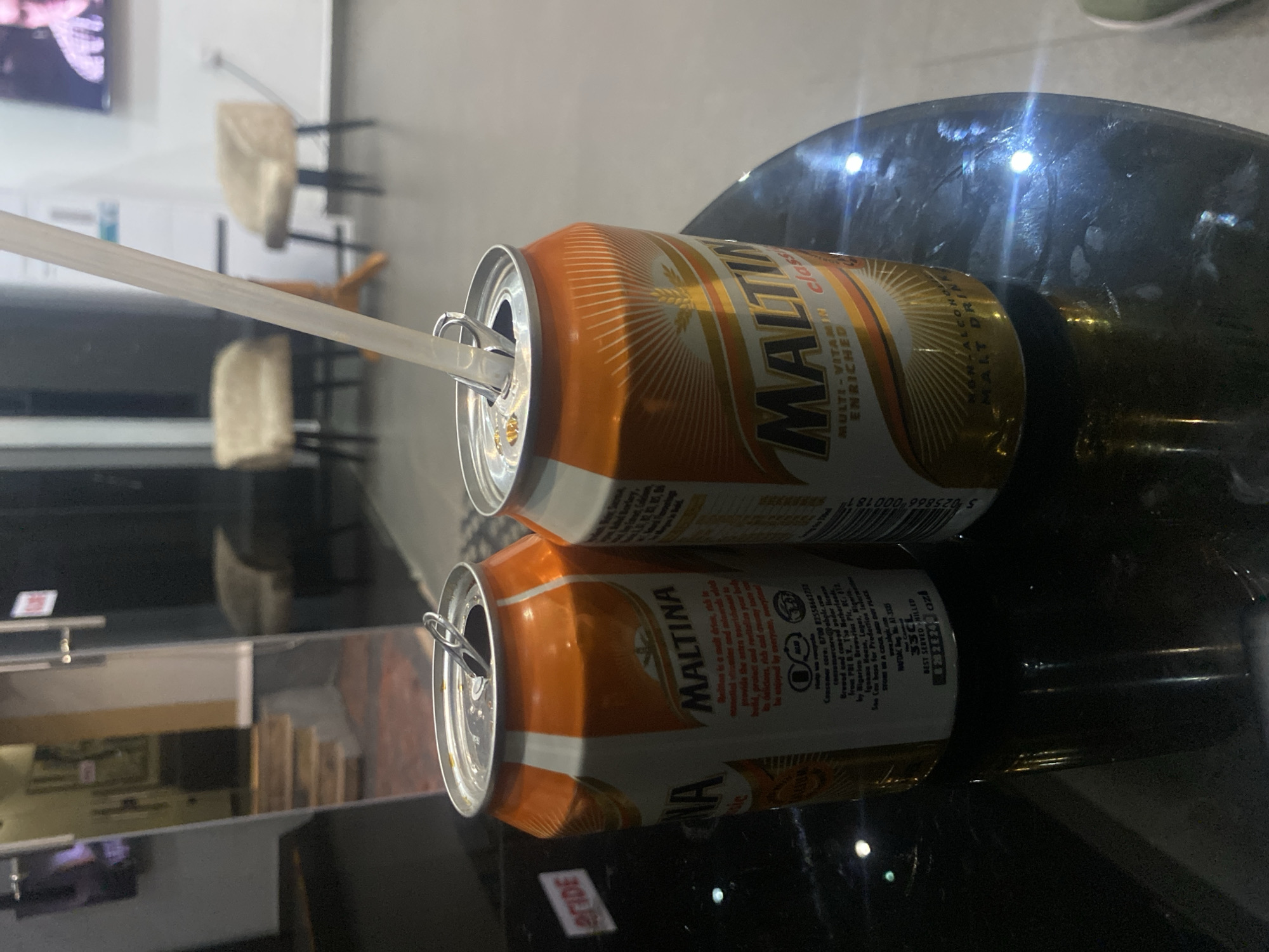 Two cans of Maltina, a non-alcoholic malt drink, are placed on a dark, reflective surface. One can is open with a straw inserted. The cans have orange and white labels with the Maltina logo and text that reads "MULTI-VITAMIN ENRICHED." In the background, there are out-of-focus chairs and a reflection of a staircase and some artwork. The lighting in the scene is dim, with reflections visible on the surface. There is no discernible information about the specific location within Barden Yana, Nigeria, or any individuals present.