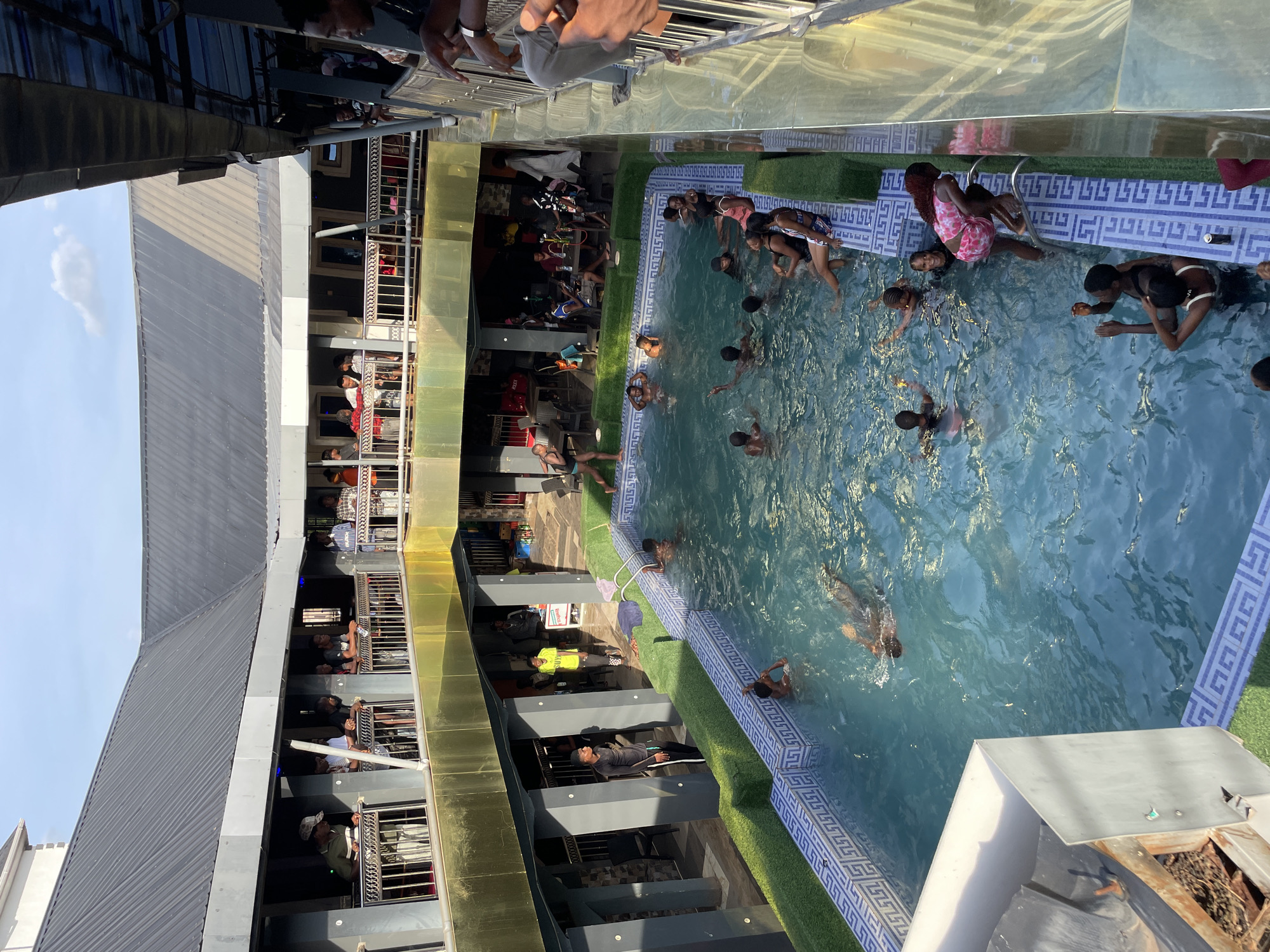 The image shows a swimming pool filled with people in Nkpor, Nigeria. The pool has a blue and white tile border with artificial grass trim. Multiple people are swimming and standing in the pool. The pool is surrounded by a multi-story building with balconies where people are watching the activity below. Some individuals are seated at tables or standing near the pool. The building has a modern architectural style with a corrugated metal roof. The sky is visible with light clouds.