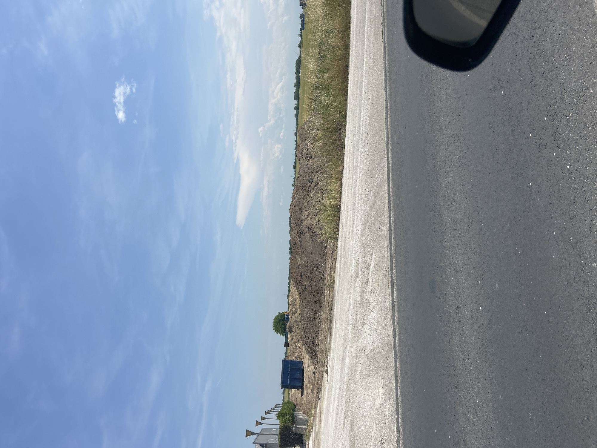 The image displays a sunny, daytime scene in Graf Ignatievo, Bulgaria, captured from a moving vehicle. The primary subjects include an asphalt road, an adjacent construction or land-clearing area, fields, and a vast blue sky.
In the immediate foreground, a dark asphalt road with a lighter-colored concrete or gravel shoulder is visible, extending from the bottom to the top right of the frame. The curved edge of a car's rearview mirror partially obstructs the upper right corner. To the left of the road, a substantial mound of dark brown, excavated soil dominates the midground. Beyond this dirt pile, a field with tall, dry grass and some greener vegetation stretches towards the horizon.
Notable objects within the scene include a prominent dark blue dumpster situated near the dirt mound and a single, lush green tree standing further back. A small, yellow piece of construction equipment, possibly an excavator or loader, is partially visible behind the dirt pile. In the distant background, faint structures and additional fields can be discerned under a bright blue sky adorned with scattered white cumulus clouds.
The weather is clear and sunny, indicative of midday or early afternoon. No people or visible text are present in the image. The setting suggests an area undergoing land development, possibly related to construction or agricultural expansion, alongside a well-maintained road.