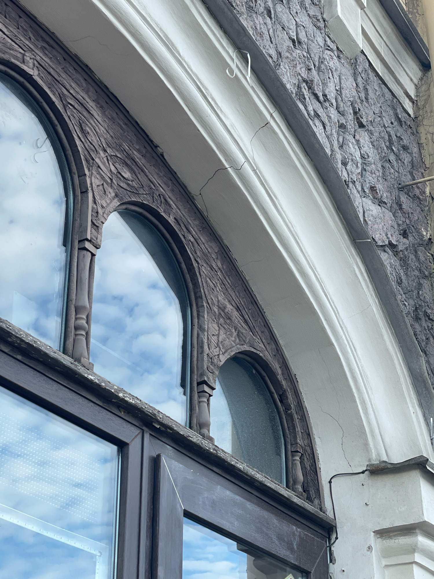 The image shows a close-up, low-angle view of the facade of a building, focusing on an arched window with dark wooden trim and intricate carvings. The window is made of large glass panes that reflect a cloudy sky. The building's exterior wall is constructed of rough, grey-toned stone blocks. Above the window, there is a curved, white architectural molding, which shows signs of cracking. The overall impression is of an older, possibly historic building. No people or specific landmarks within Saint Petersburg are visible.
