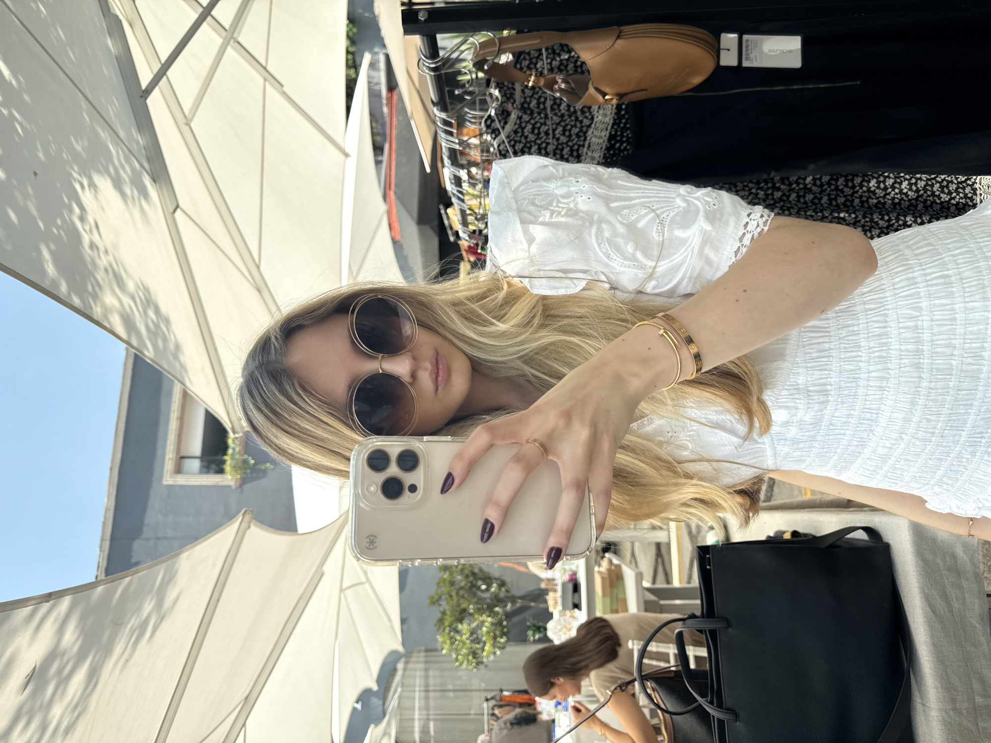 A blonde woman takes a selfie in Mexico City. She is wearing sunglasses and a white dress, with gold bracelets on her left wrist. She holds a beige iPhone in her hand, showcasing dark purple manicured nails. Behind her is an umbrella and clothing rack with handbags. In the foreground, another woman is seated. A black tote bag is on the table. The outdoor setting appears to be a marketplace.