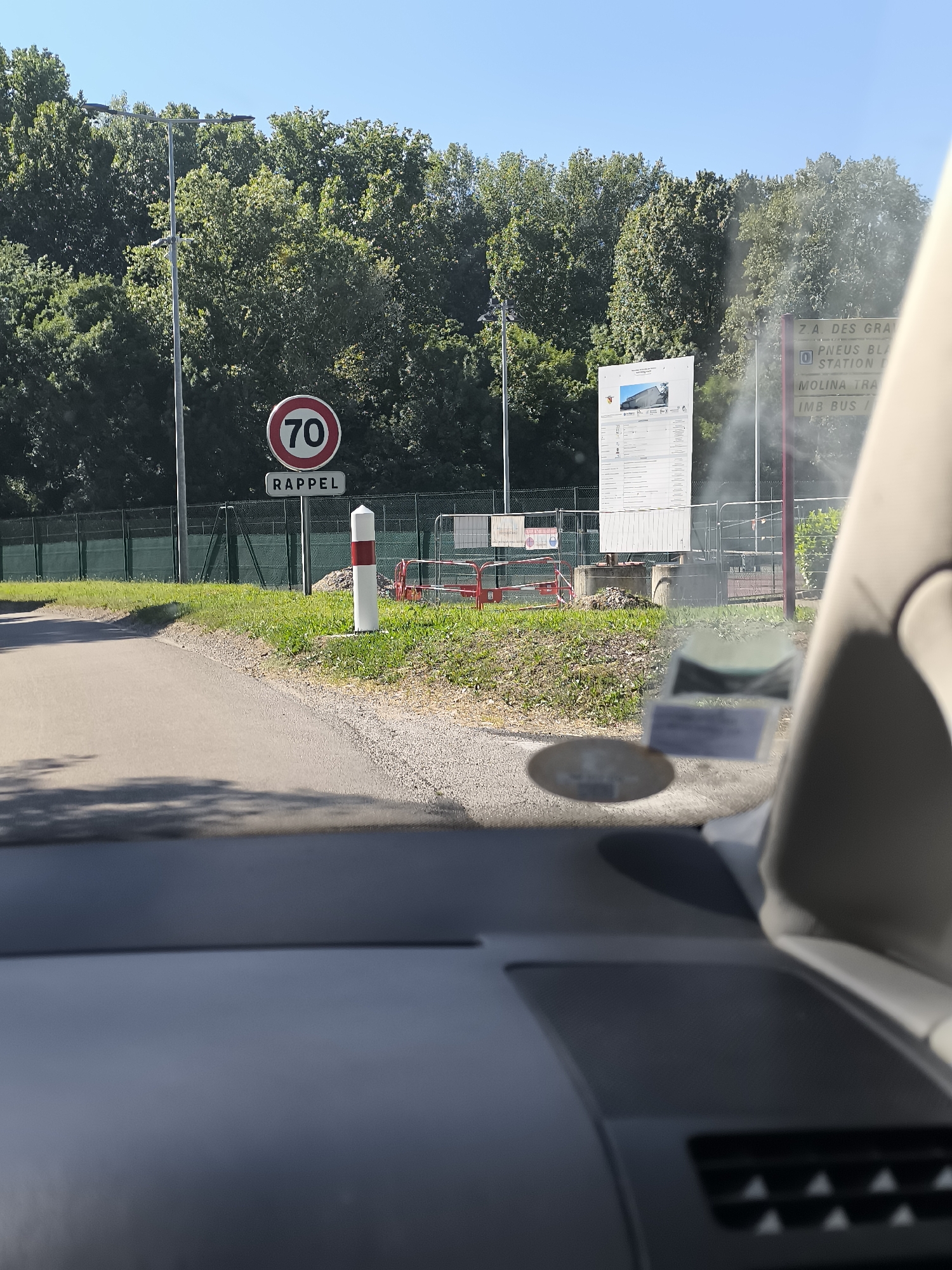 The image depicts a roadside scene in Saint-Pierre-de-Bœuf, France, viewed from inside a vehicle. Visible outside the vehicle are a speed limit sign indicating "70 RAPPEL" (reminder of a 70 km/h speed limit), a red and white striped bollard, and a large sign displaying project information near an active construction site. The sign appears to be related to the nearby "Z.A. DES GRANDS" industrial zone, as evidenced by the visible text "Z.A. DES GRANDS" and names of businesses or locations like "PNEUS BLAN", "MOLINA TRA", and "IMB BUS". A green fence runs along the perimeter, with trees forming a dense backdrop. The foreground is the dashboard of a vehicle with a registration sticker. The scene is bathed in daylight, under a clear sky.