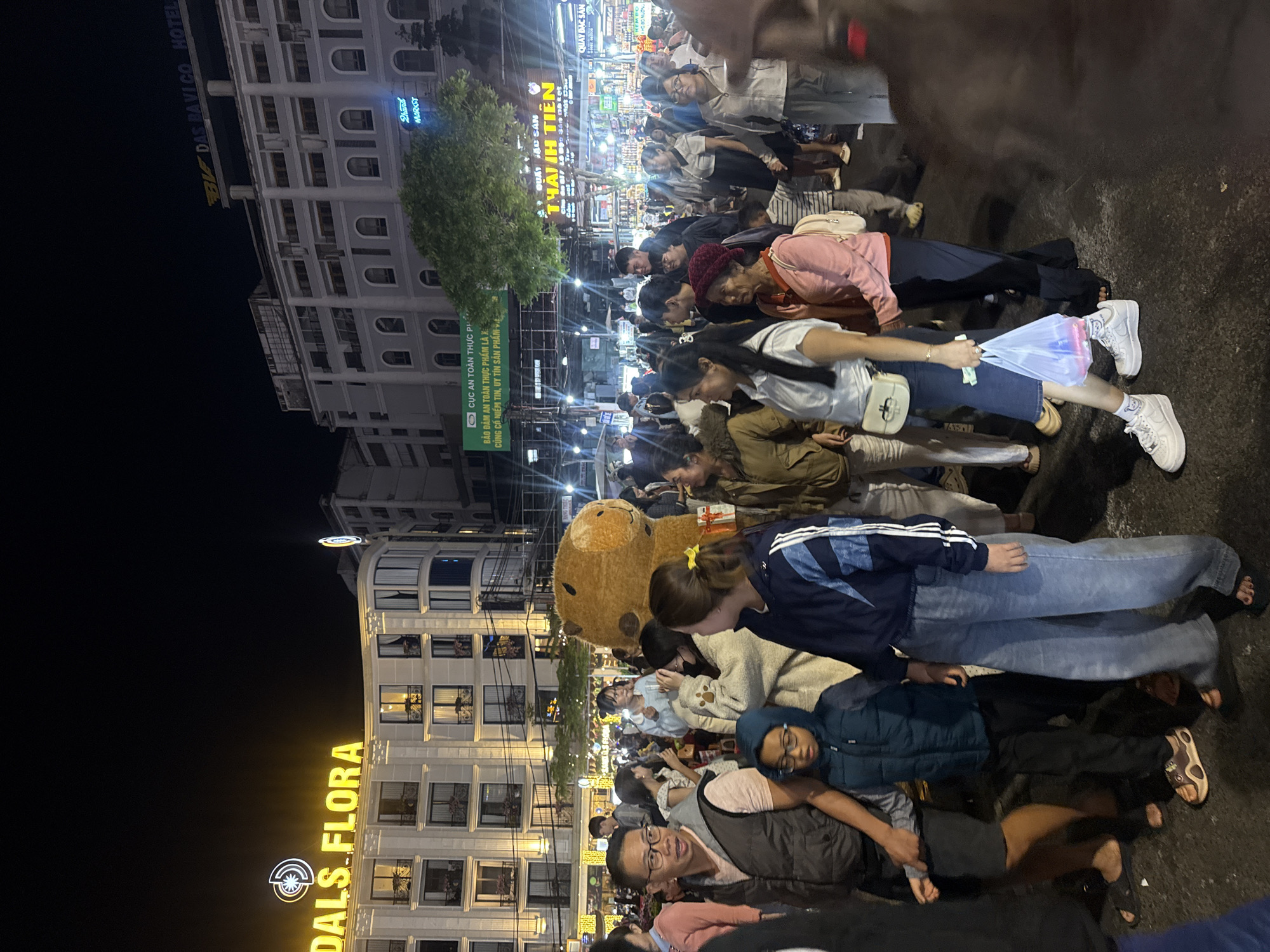 The image depicts a nighttime street scene in Da Lat, Vietnam, likely a bustling market or pedestrian area. In the background, illuminated buildings display prominent signage. One building has a glowing yellow sign that reads "DALS.FLORA" with a circular logo above it. Another building in the distance features a yellow sign with a stylized "BF" logo and the text "DAS PAVICO HOTEL". A green banner with Vietnamese text is also visible across a street.
The foreground and midground are filled with a crowd of people of various ages, dressed in casual, layered clothing suitable for a cooler evening. Many individuals are walking, some in pairs or small groups, suggesting a social gathering or a shopping excursion. A large, light-brown, teddy bear mascot costume is a central figure in the midground, with a person inside wearing it. People are interacting with each other, looking in different directions, and some are holding bags. The ground appears to be asphalt, wet in places, reflecting the ambient light. The overall atmosphere is lively and illuminated by artificial lights from buildings and street vendors.