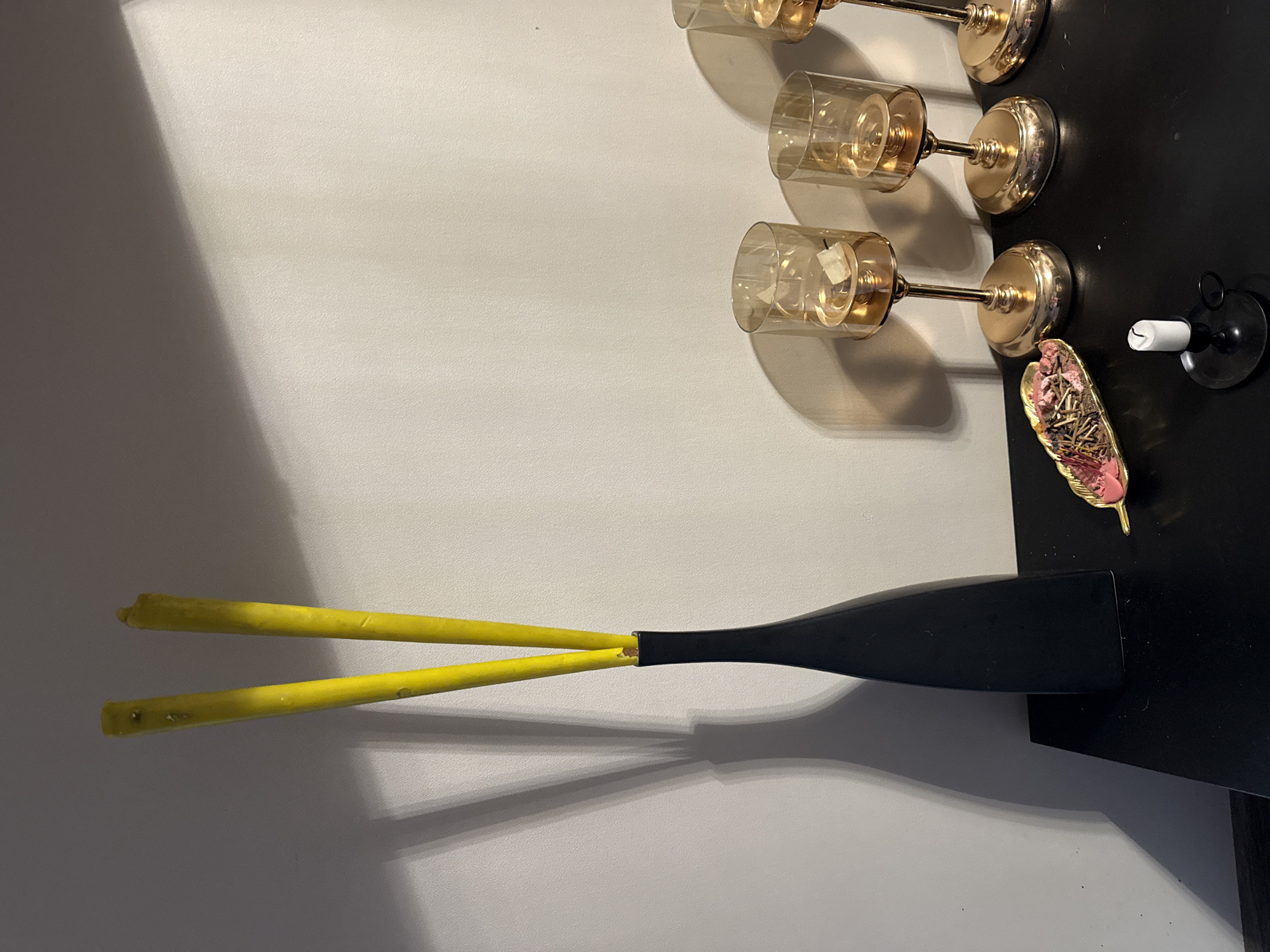 In an indoor setting in Berezyna, Ukraine, a dark, flat surface, likely a table or shelf, is positioned against a light-colored wall. Strong, directional lighting from an unseen source casts elongated shadows across the wall and the table.
On the left side of the table, a tall, slender black vase holds two bright yellow, thin, rod-like objects, possibly candles or decorative sticks. These objects and the vase cast prominent, elongated shadows onto the wall behind them, extending upwards and to the left. The yellow objects show some minor surface imperfections or marks, particularly near their tips.
Towards the right side of the table, three golden-colored candle holders are arranged in a horizontal line. Each holder features a clear glass cylinder atop a golden stem and a rounded base. The leftmost glass cylinder contains a small, light-colored, amorphous object. The other two cylinders appear empty.
In the foreground, to the left of the golden candle holders, a small, leaf-shaped, golden-colored dish contains an assortment of dried organic material, including small sticks and pink-colored elements, suggestive of potpourri. Directly in front of this dish, a small, white cylindrical candle sits in a black, circular candle holder with a small handle.
No individuals are present, and no actions or interactions are depicted in the scene.