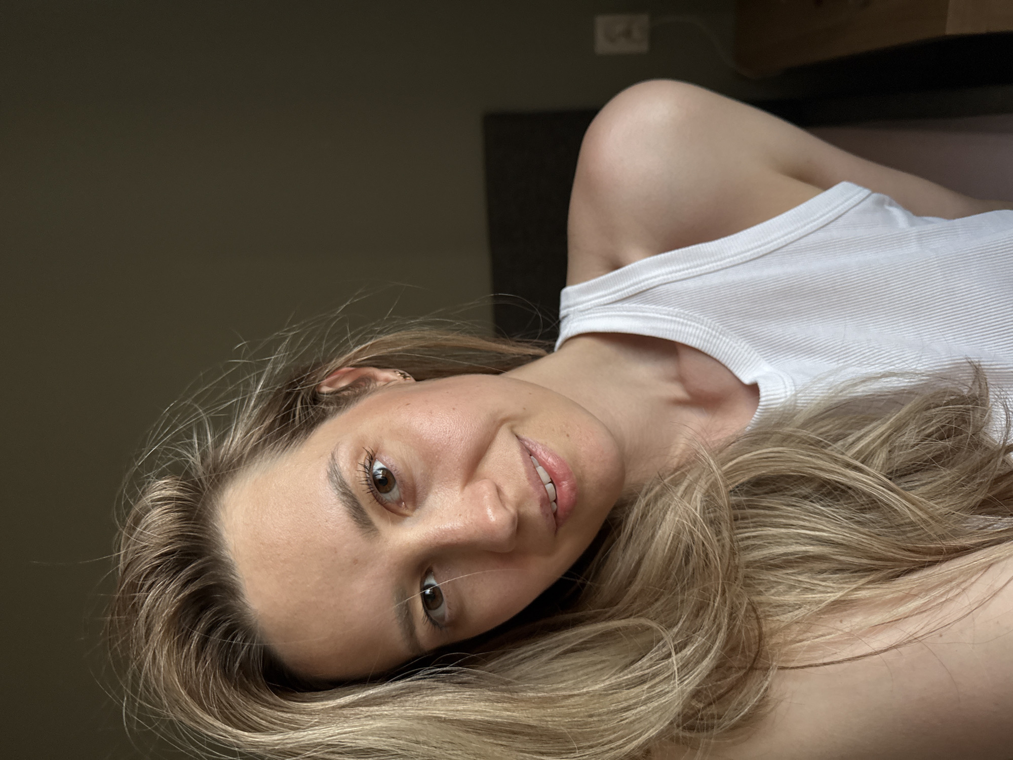 A woman with long, light-brown hair is lying on her side, wearing a white tank top. The background consists of a dark wall and a partially visible wooden cabinet. The location is not possible to determine from the image.