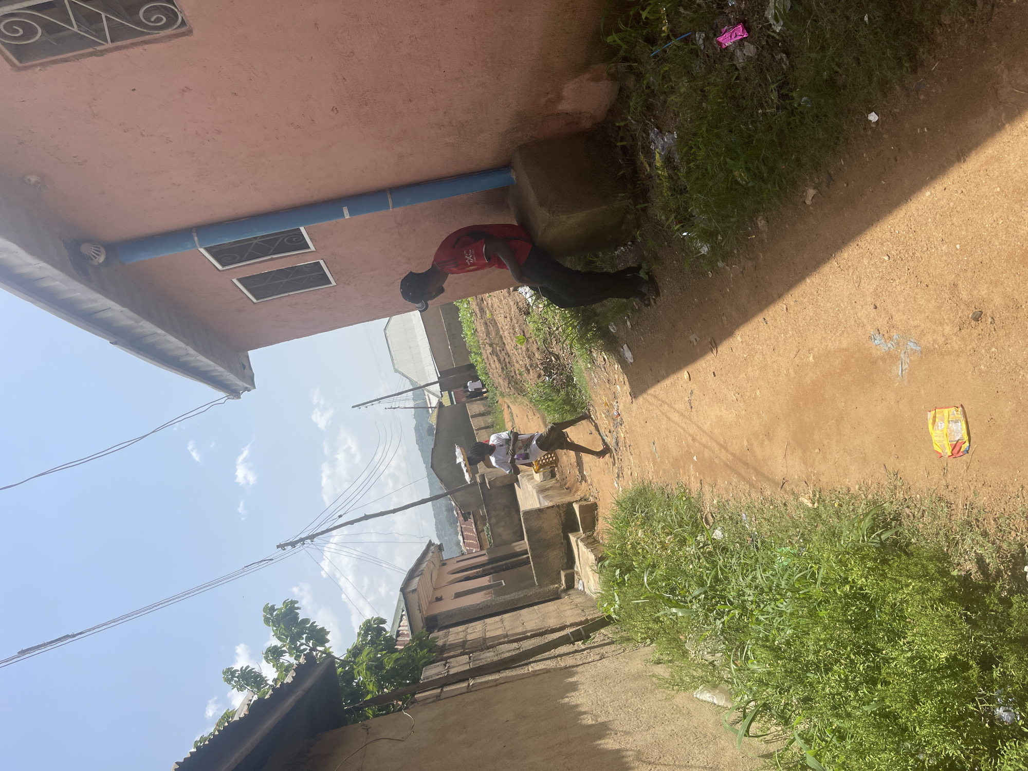 The image, taken in Rafin Par, Nigeria, depicts an outdoor scene under a clear sky, suggesting daytime. The perspective is from ground level, looking upwards and slightly sideways.
The main subjects are two young males. One, wearing a red shirt and black shorts, is leaning against a concrete structure, facing away from the camera. Another boy, dressed in a white t-shirt and shorts, is standing further down a dirt path. He appears to be holding something in his hands, possibly a basket, and looking towards the ground.
The setting is an urban or semi-urban environment, characterized by buildings with concrete and reddish-pink walls. There are visible electrical wires suspended on poles, indicating infrastructure. A blue pipe runs along the exterior of one building. The ground is a mixture of dirt and some vegetation, with scattered litter visible, including a brightly colored snack wrapper. The overall impression is of a modestly developed area.
The image lacks strong emotional cues, but the casual posture of the first boy and the focused stance of the second suggest everyday activities. There is no visible text in the image. The lighting suggests it is a sunny day.
