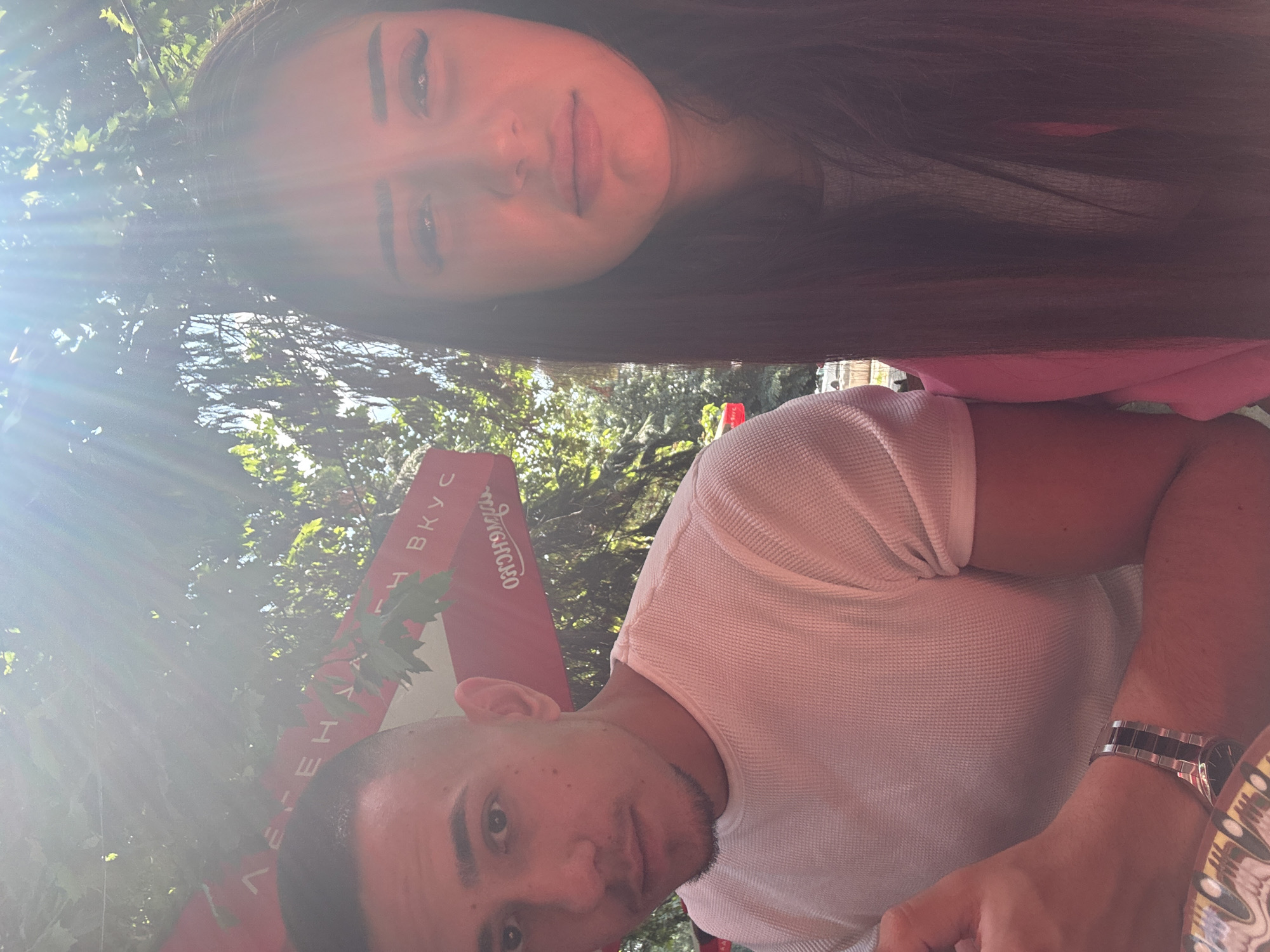This image captures two individuals posing outdoors on a sunny day, likely in a cafe or restaurant setting in Plovdiv, Bulgaria. The photo is oriented ninety degrees counter-clockwise from a standard upright perspective.
In the upper portion of the frame (when correctly oriented), a woman with long, dark hair is visible, looking towards the camera. She has light skin and a calm, slightly contemplative expression with softly pursed lips. She appears to be wearing a pink top. Below her, occupying the lower part of the frame, is a man also looking towards the camera. He has short dark hair and light stubble on his chin and upper lip, with a neutral to slightly smirking expression. He is wearing a white textured (waffle-knit) t-shirt, and a metallic watch with a black and silver band is visible on his left wrist.
The background features lush green foliage, suggesting an outdoor location with trees or bushes. A prominent sun flare or lens flare emanates from the upper left of the image (when correctly oriented), casting a bright glow that creates a vibrant, summery atmosphere. Between the two individuals, a red banner or sign is partially visible, displaying text in Cyrillic characters. Clearly legible words include "ВКУС" (VKUS, meaning "taste") and "ЛЕГЕНДА" (LEGENDA, meaning "legend"), along with fragments of other words like "СОУСОВ" (SOUSOV, meaning "sauces"). The overall scene conveys a casual, relaxed moment captured in bright daylight.