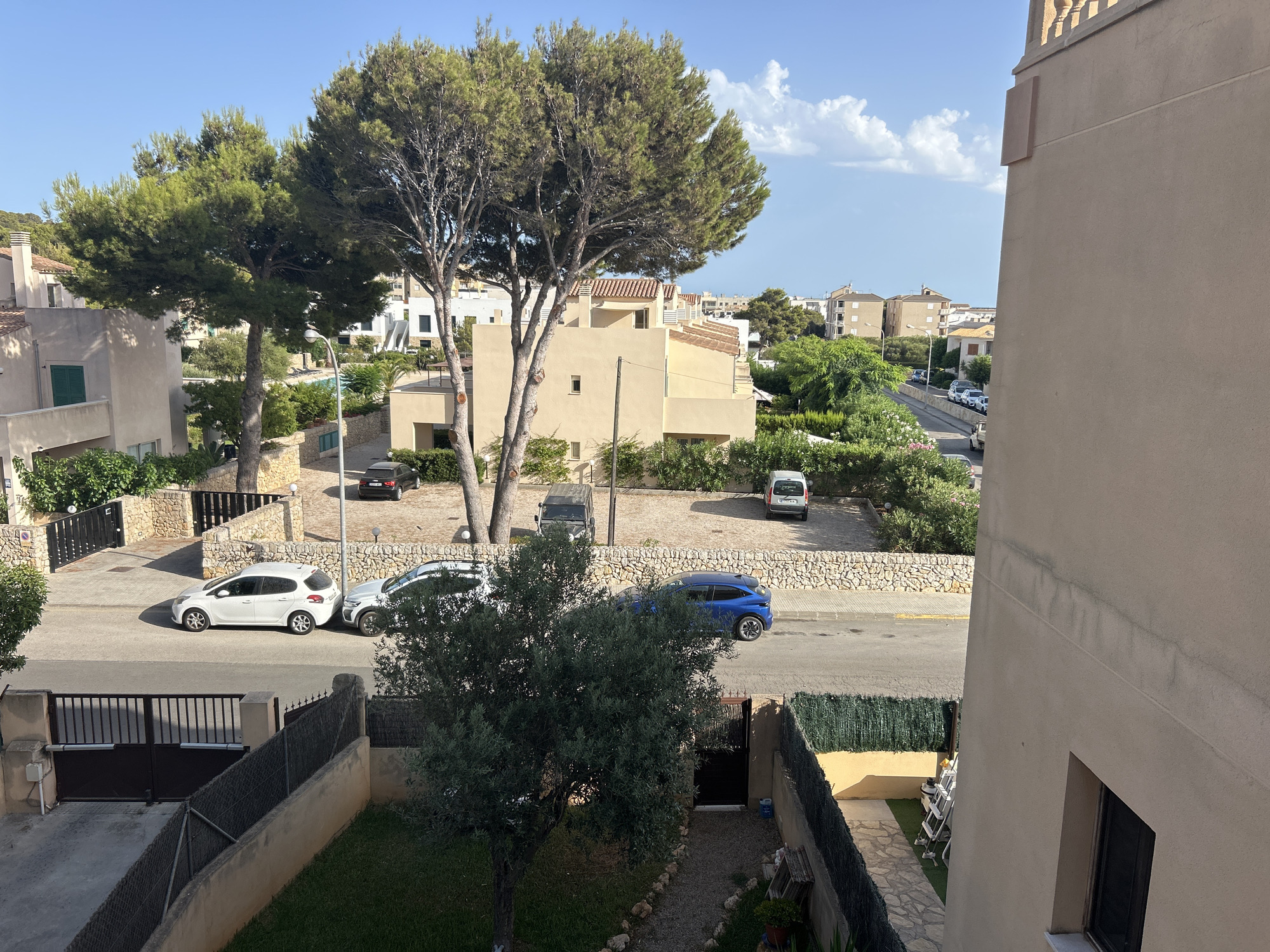 This is a photograph taken from an elevated vantage point, looking down onto a street in Capdepera, Spain. The scene depicts a residential area with multiple buildings, trees, and parked cars. On the right side of the image, a large, light-beige building dominates the foreground, with a window visible. To the left of this building, a street runs horizontally across the frame. Several cars are parked along the street, including a white hatchback, a silver SUV, and a blue SUV. Beyond the street, there is a stone wall, followed by more buildings and lush green trees, including two large pine trees in the middle ground. In the background, more houses and buildings are visible, suggesting a densely populated urban environment, with a clear blue sky above, dotted with a few white clouds. The overall impression is one of a sunny day in a Mediterranean town.